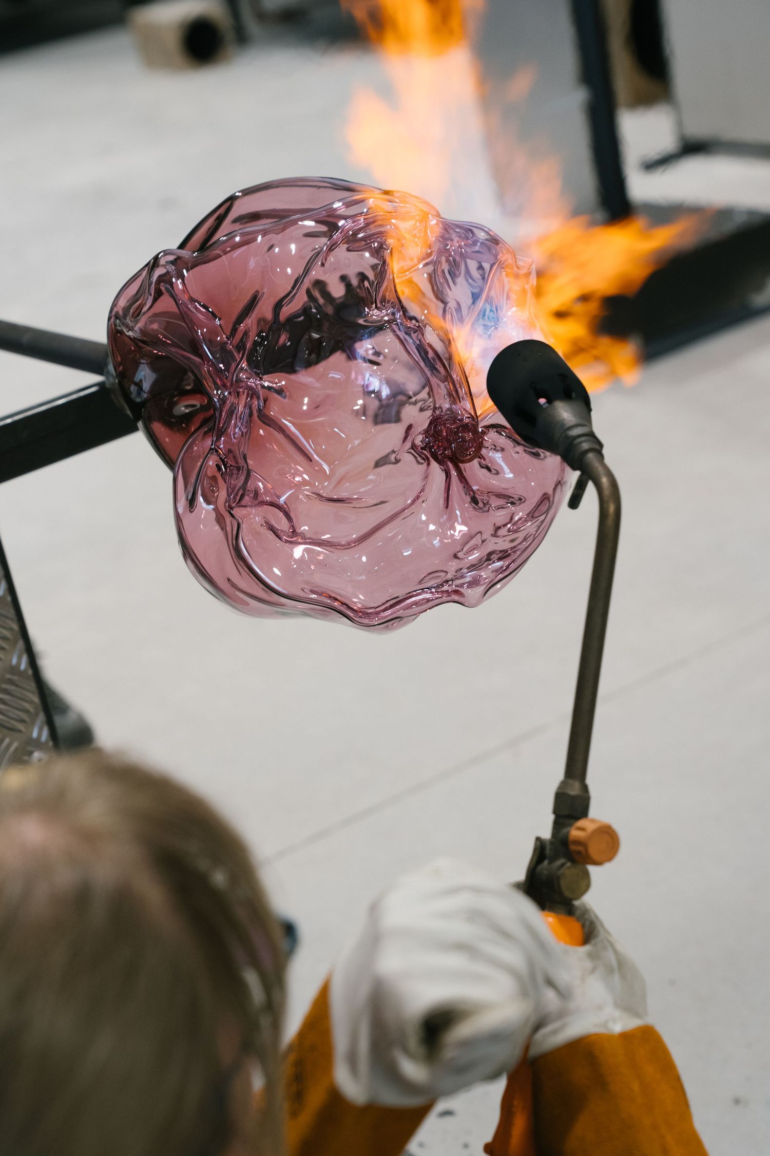 Glassblower in protective gloves heats a delicate pink glass piece with a blowtorch flame in a studio.