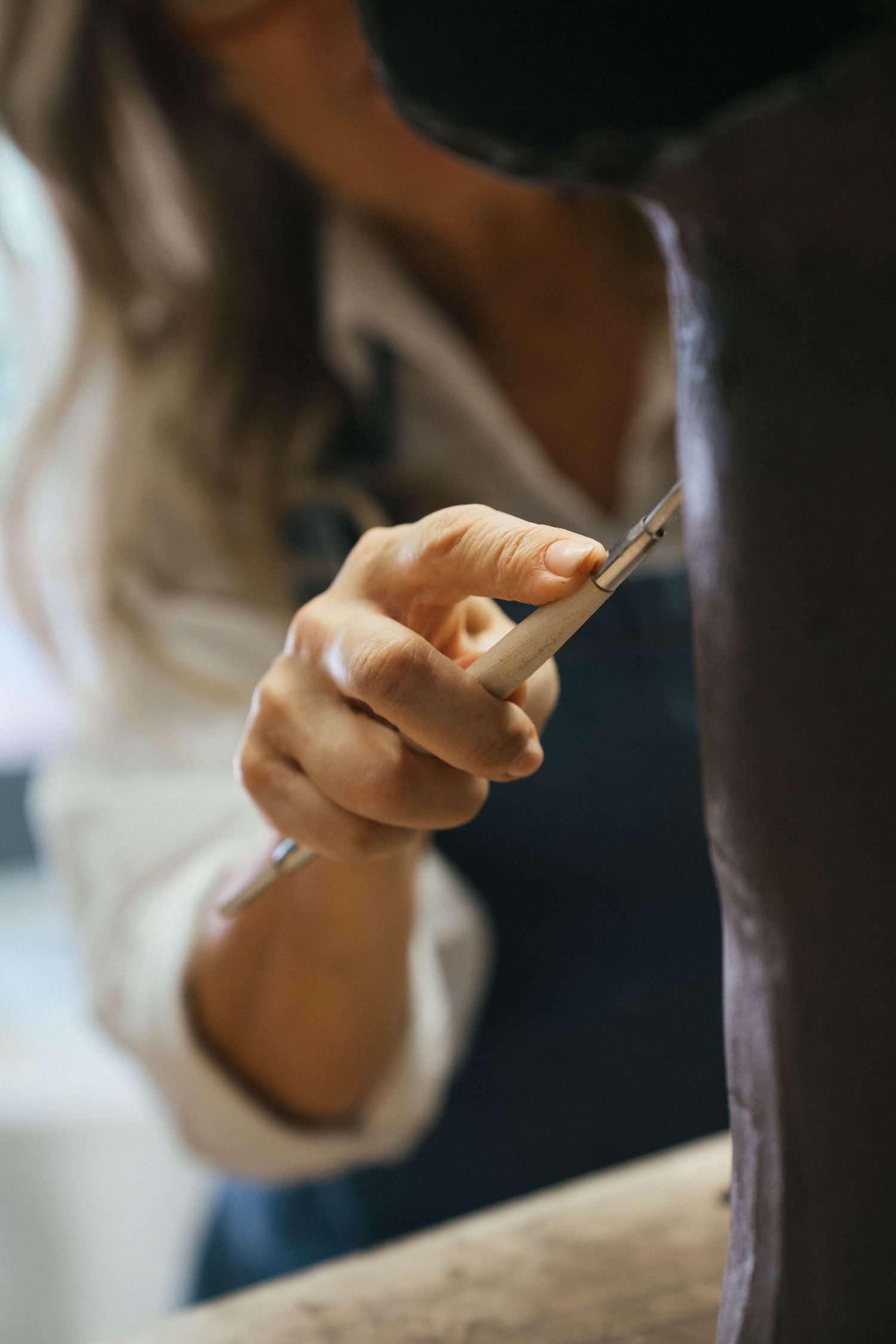 Artisan's hand uses a fine sculpting tool to carve detail into a dark clay ceramic piece in a workshop.