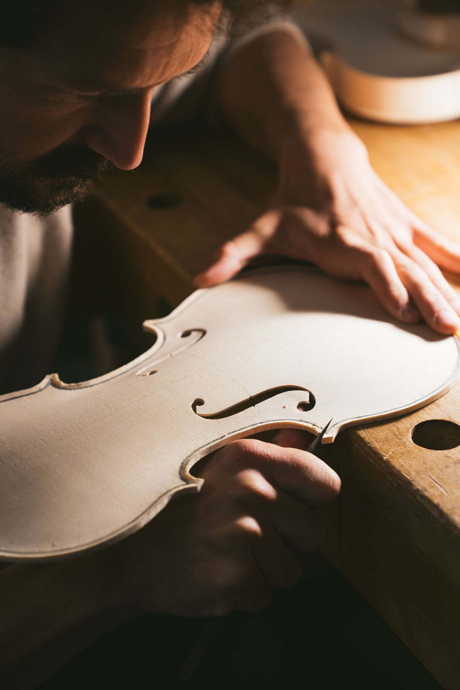 A violin maker leans over a raw maple and spruce violin body, inspecting the f-holes and edges at a wooden workbench in a workshop.