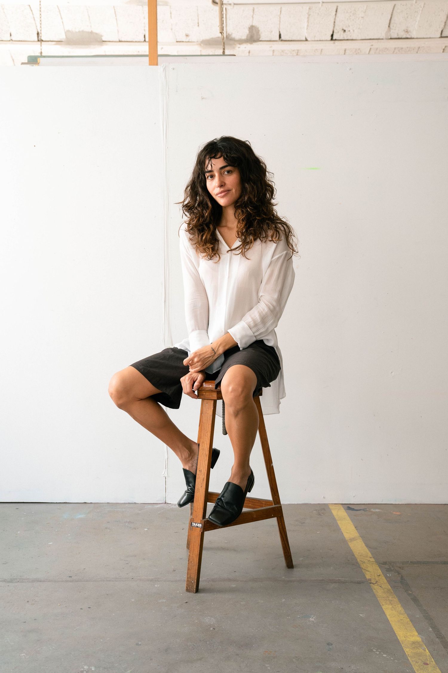 Woman with curly dark hair, white blouse and black shorts sits on a wooden stool in a bright studio with a white wall.