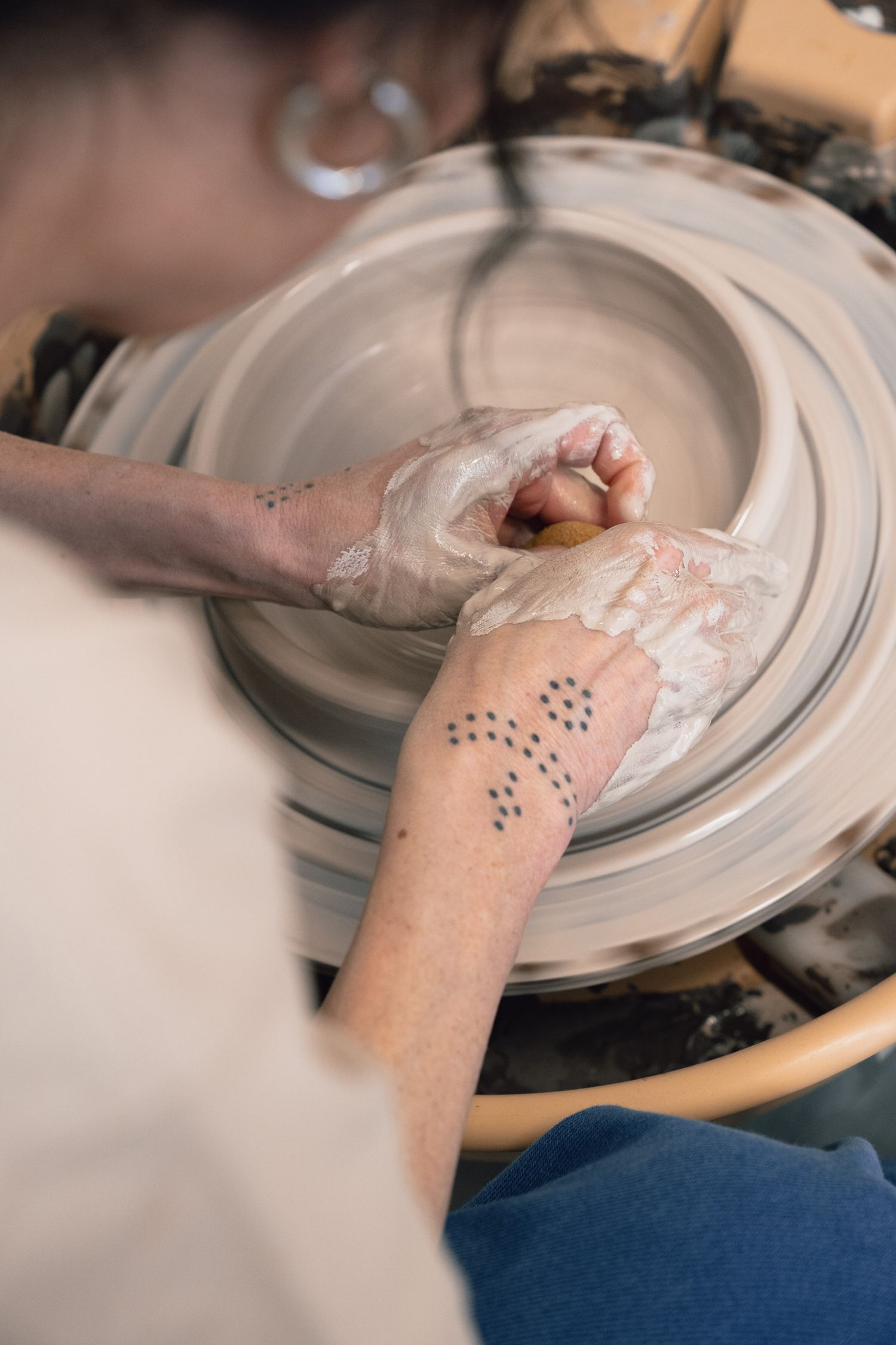 Potter with clay-covered hands shaping a bowl on a spinning pottery wheel, wearing a hoop earring and dotted hand tattoo.