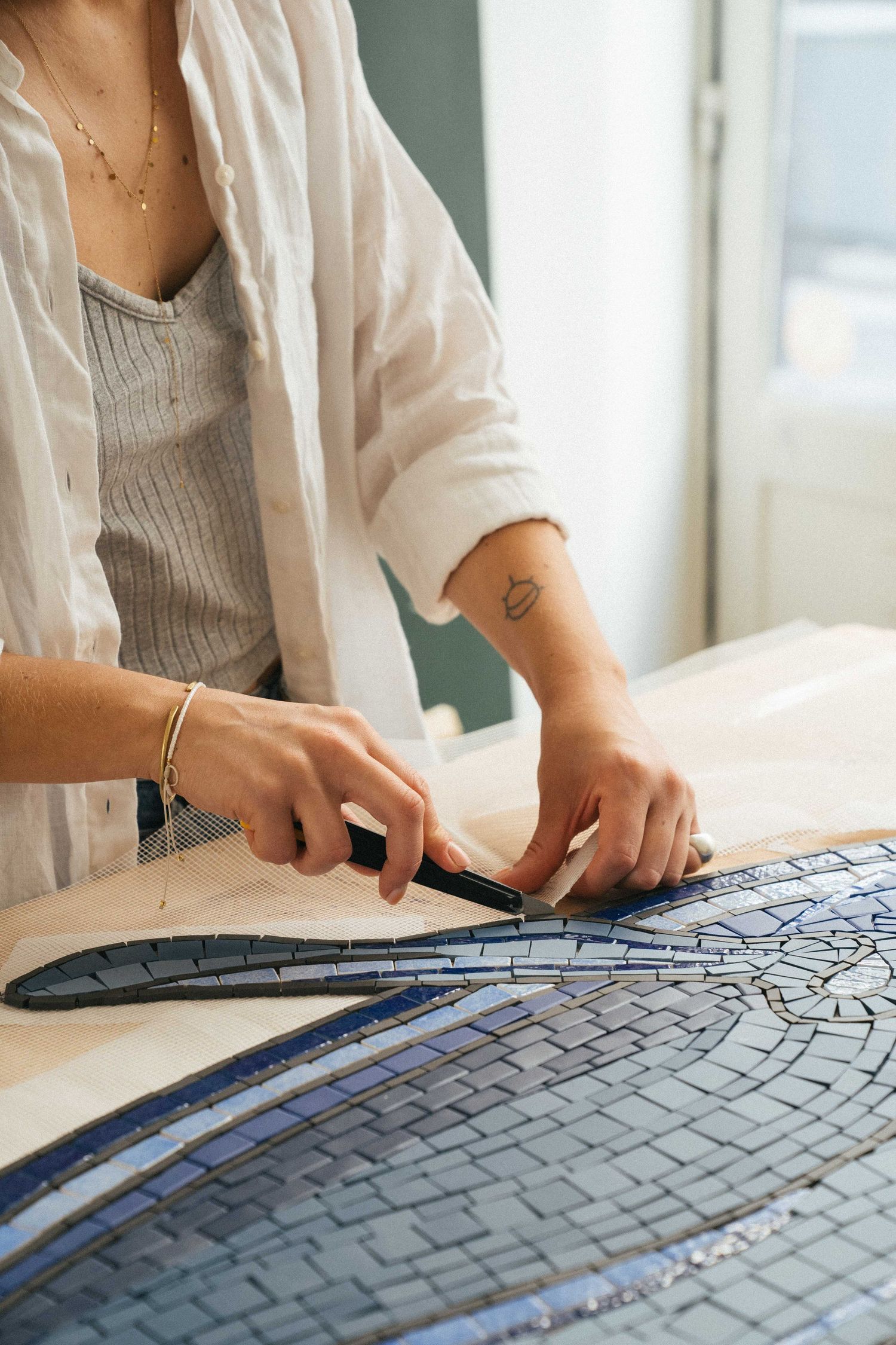 Artisan in a white linen shirt cuts and arranges small blue tiles to create a detailed mosaic on a worktable.