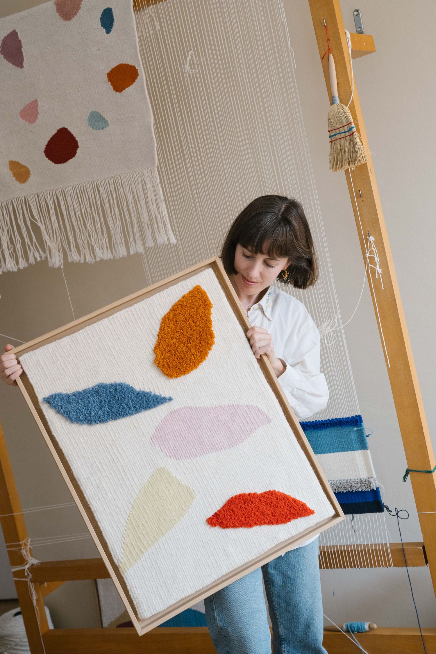 Woman holding a framed handwoven tufted wall hanging with colorful organic shapes in front of a loom in a weaving studio.