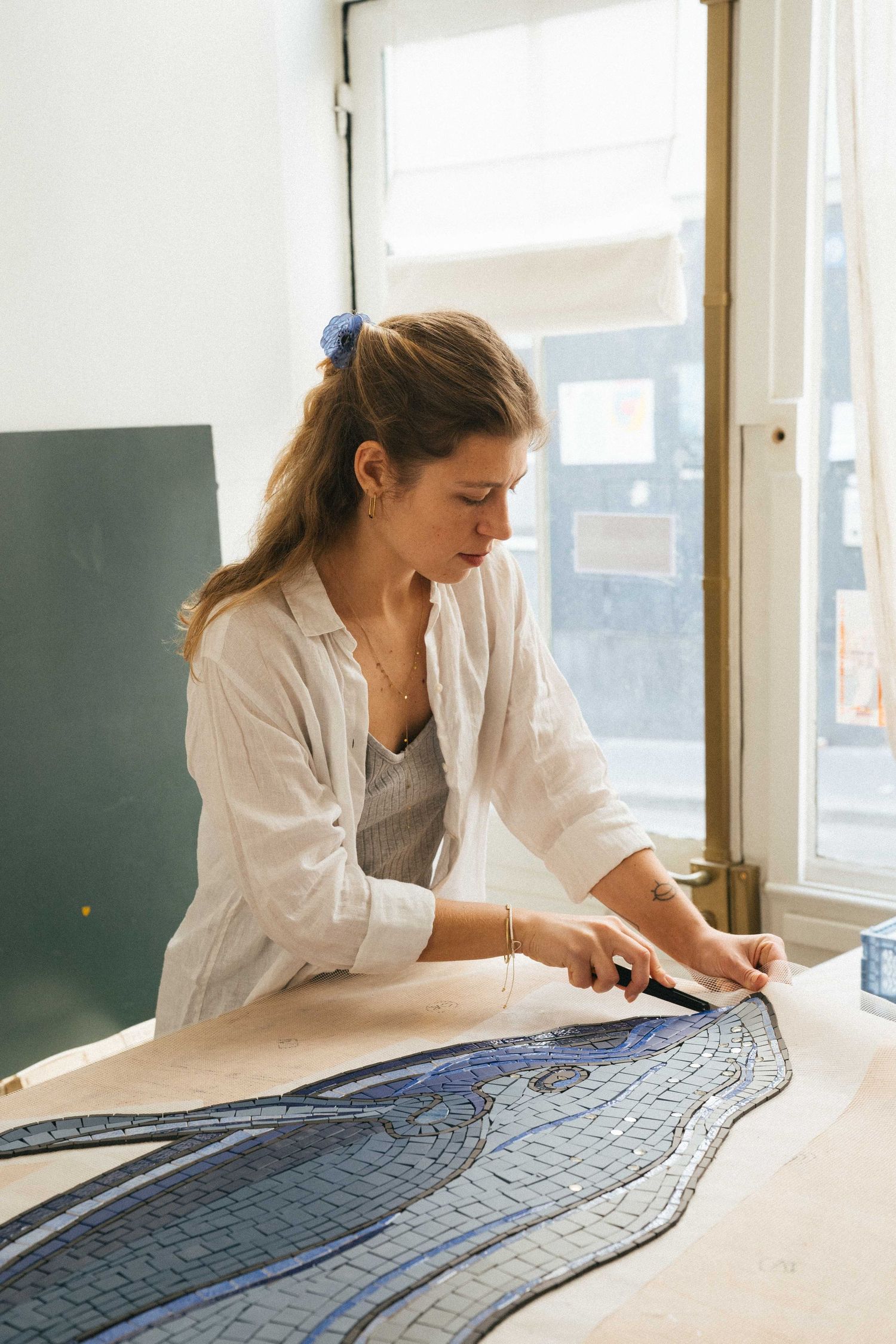 Artist with a blue hair tie arranges small blue and gray tiles on a wavy mosaic design at a worktable beside a bright studio window.