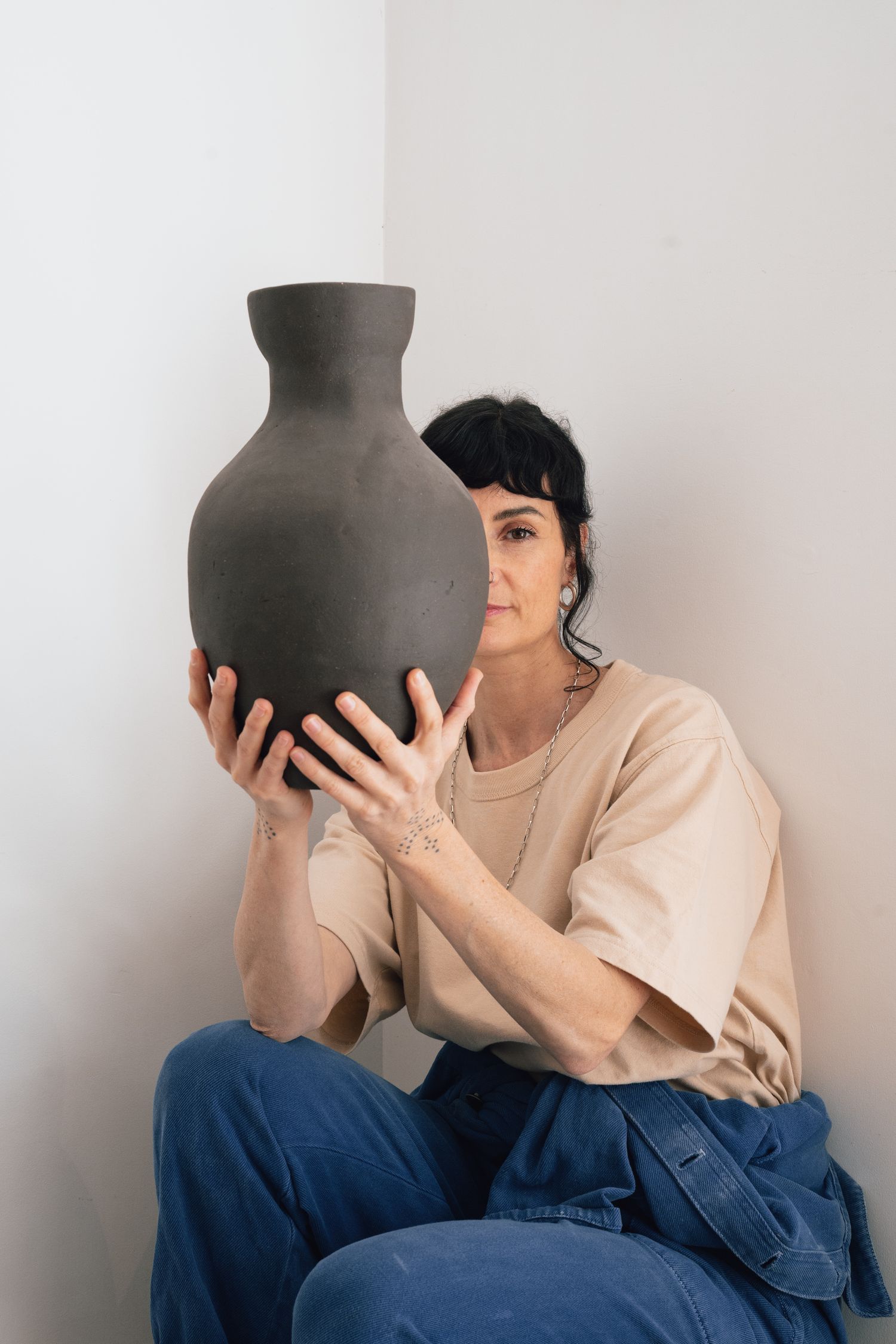 Woman in a beige t-shirt and blue jeans sits in a corner holding a large handmade dark gray ceramic vase in front of her face.