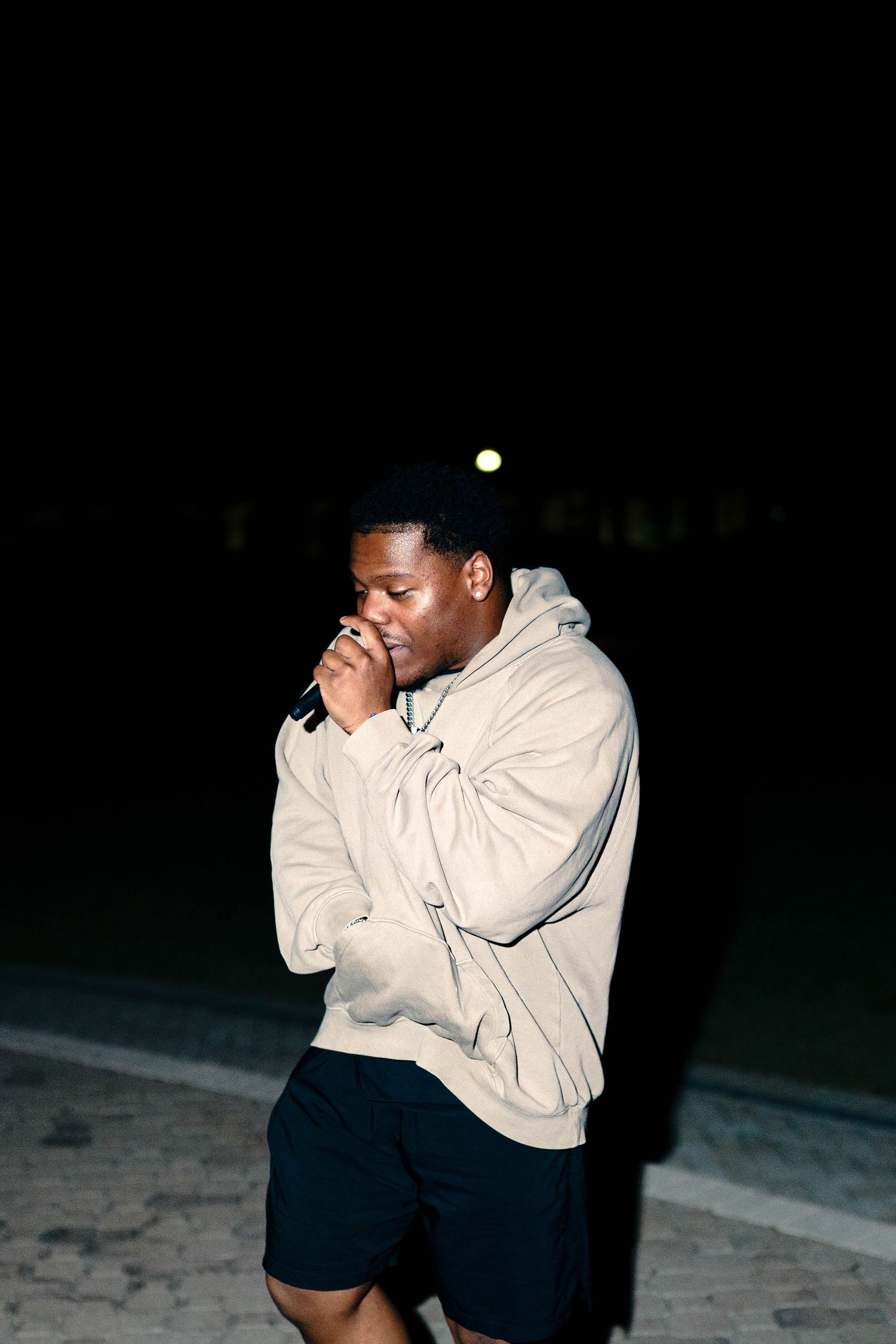 Young Black man in beige hoodie and black shorts laughing outdoors at night under dim lighting.