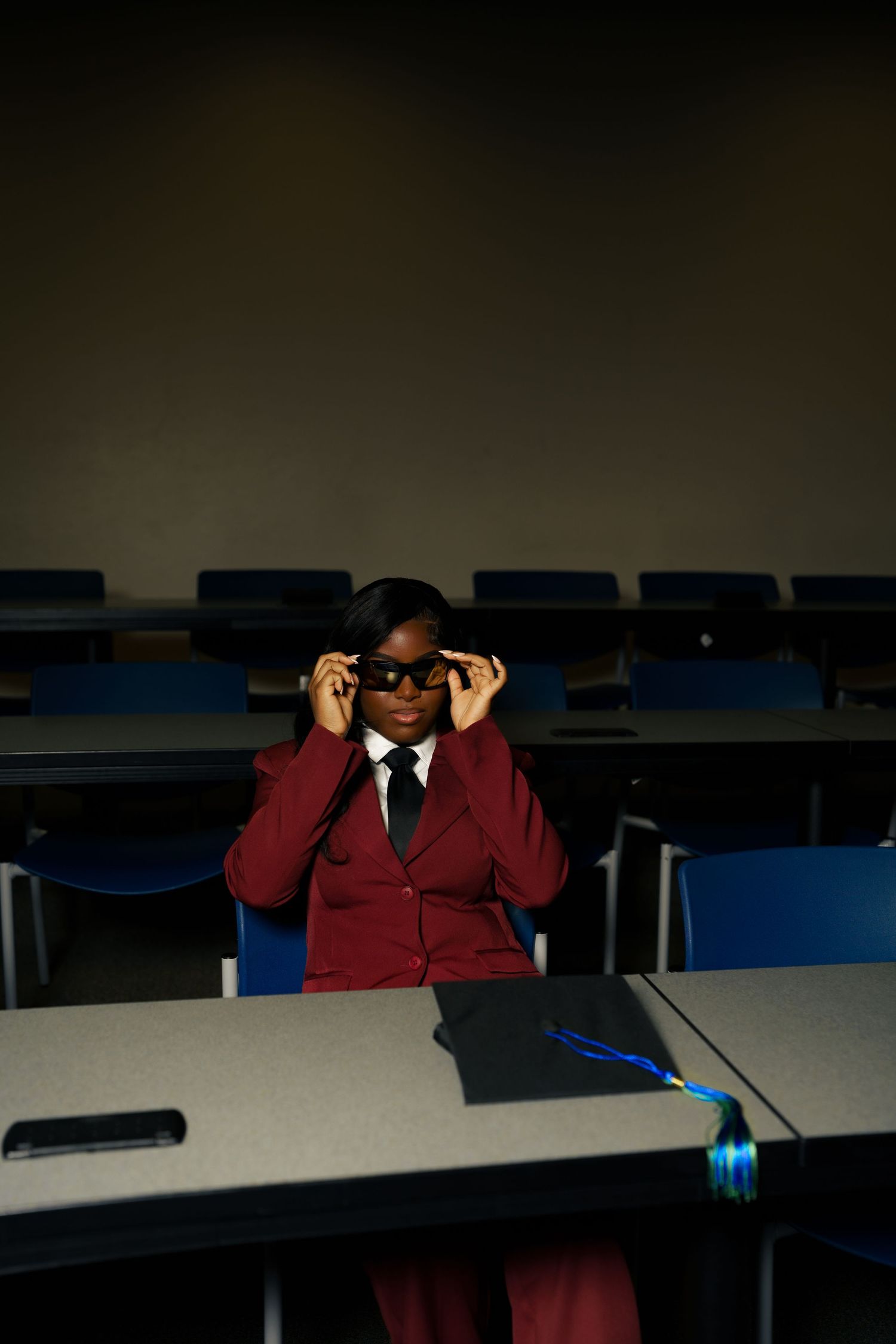 A woman in a red suit adjusts her glasses while seated in an empty lecture hall with a graduation cap on the desk.