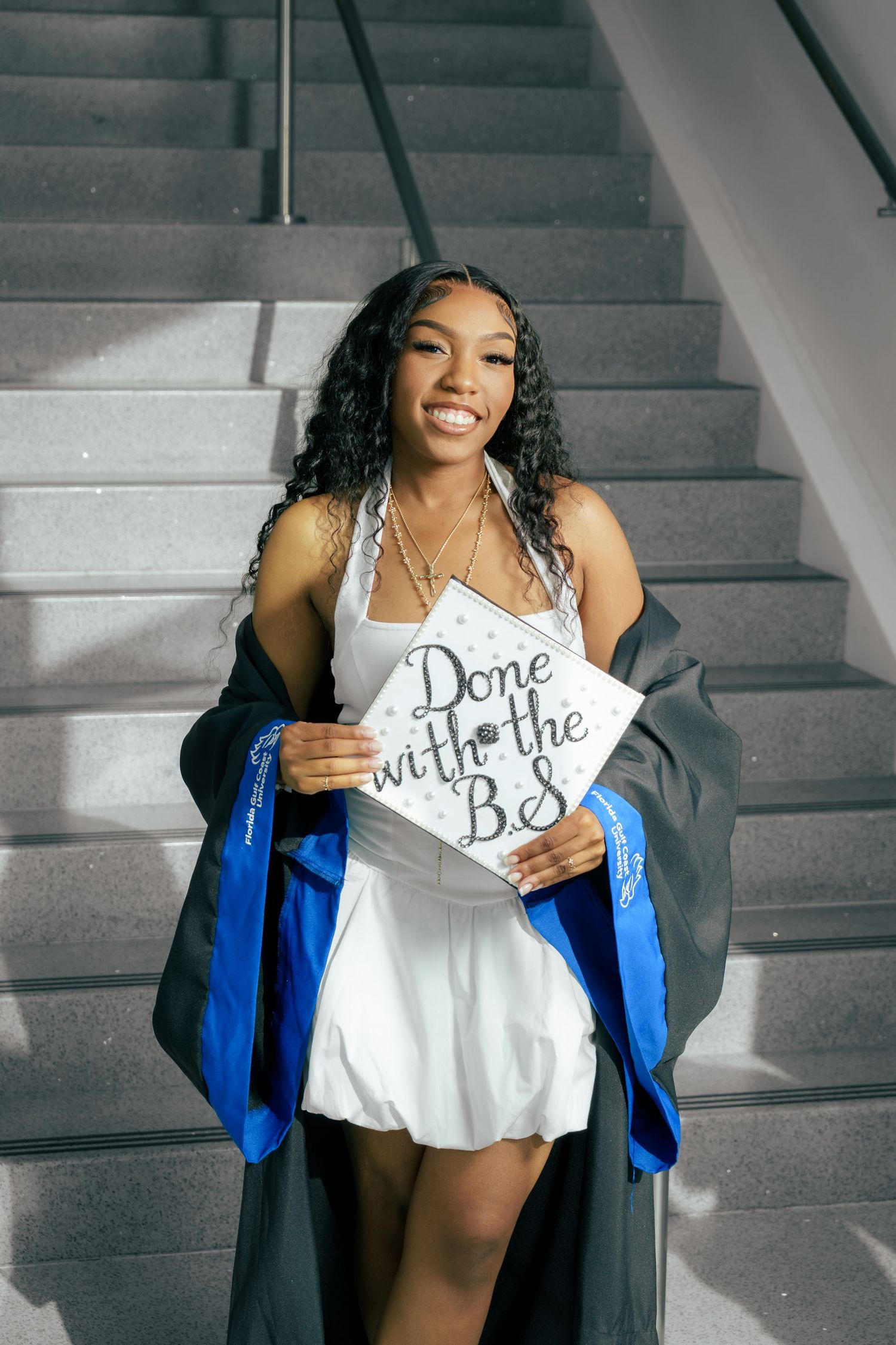 Graduate in white dress and graduation gown holds a decorative 'Done with the Degree' sign on concrete stairs.