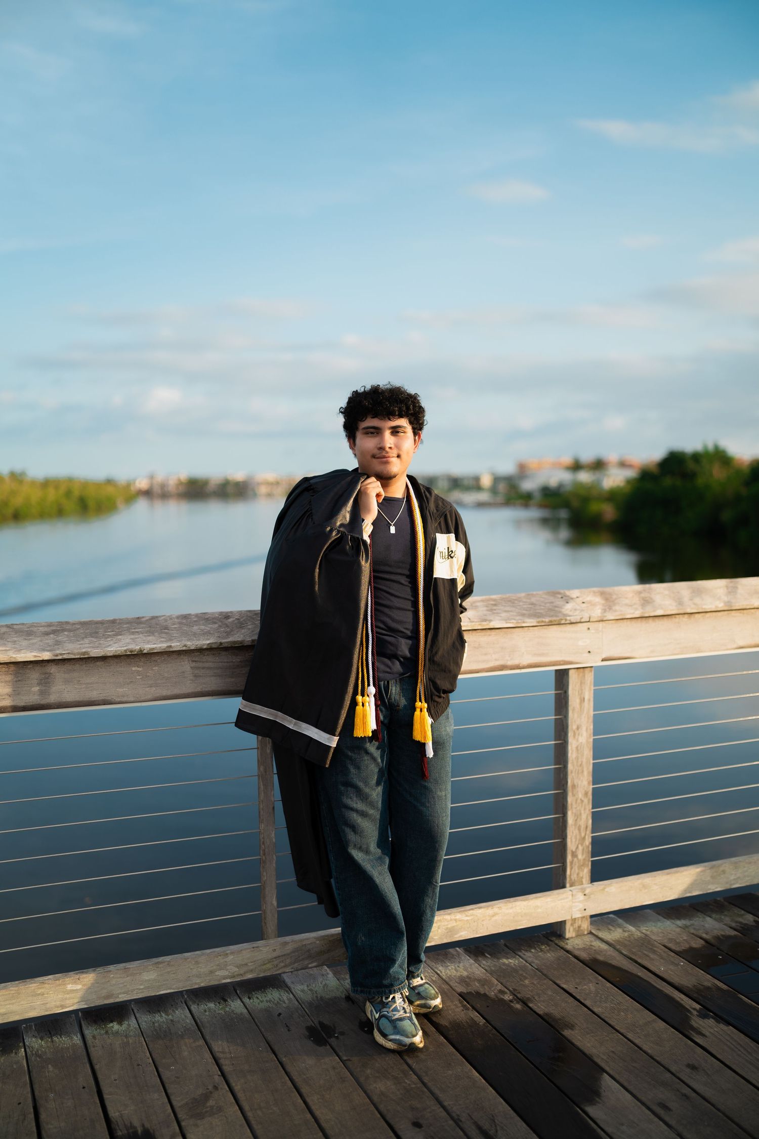 Graduate in cap and gown holds tassel on wooden bridge overlooking calm water at golden hour.
