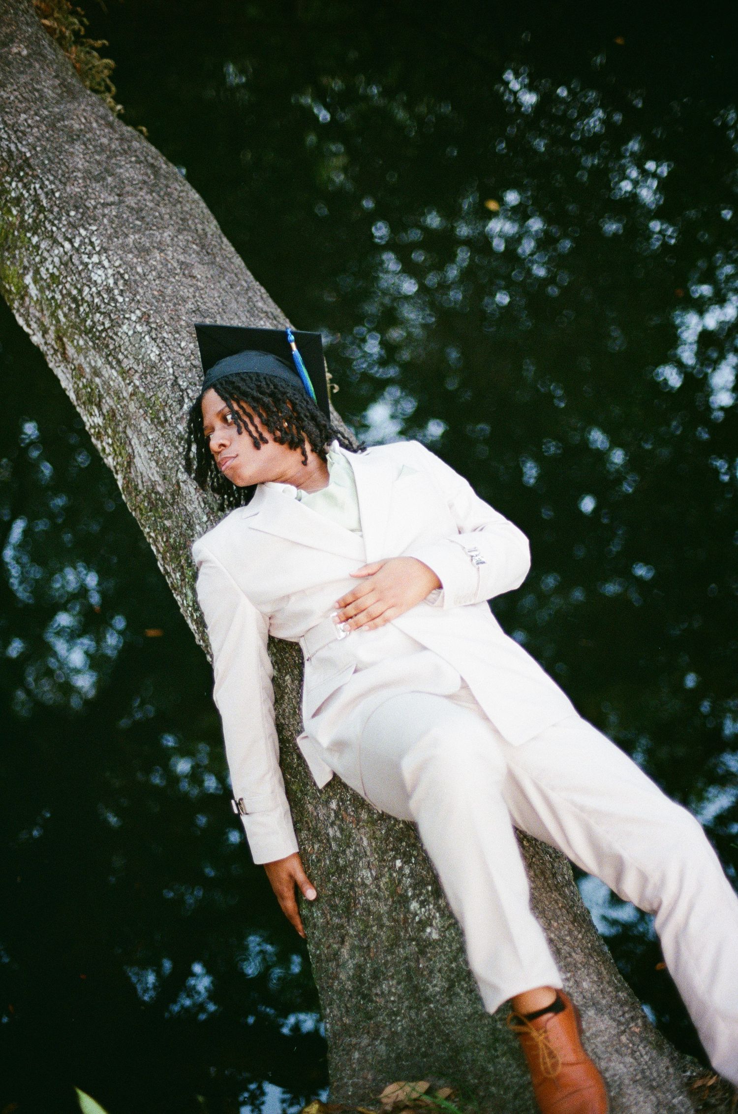 A graduate in a white suit and cap reclines gracefully against a tree trunk outdoors.