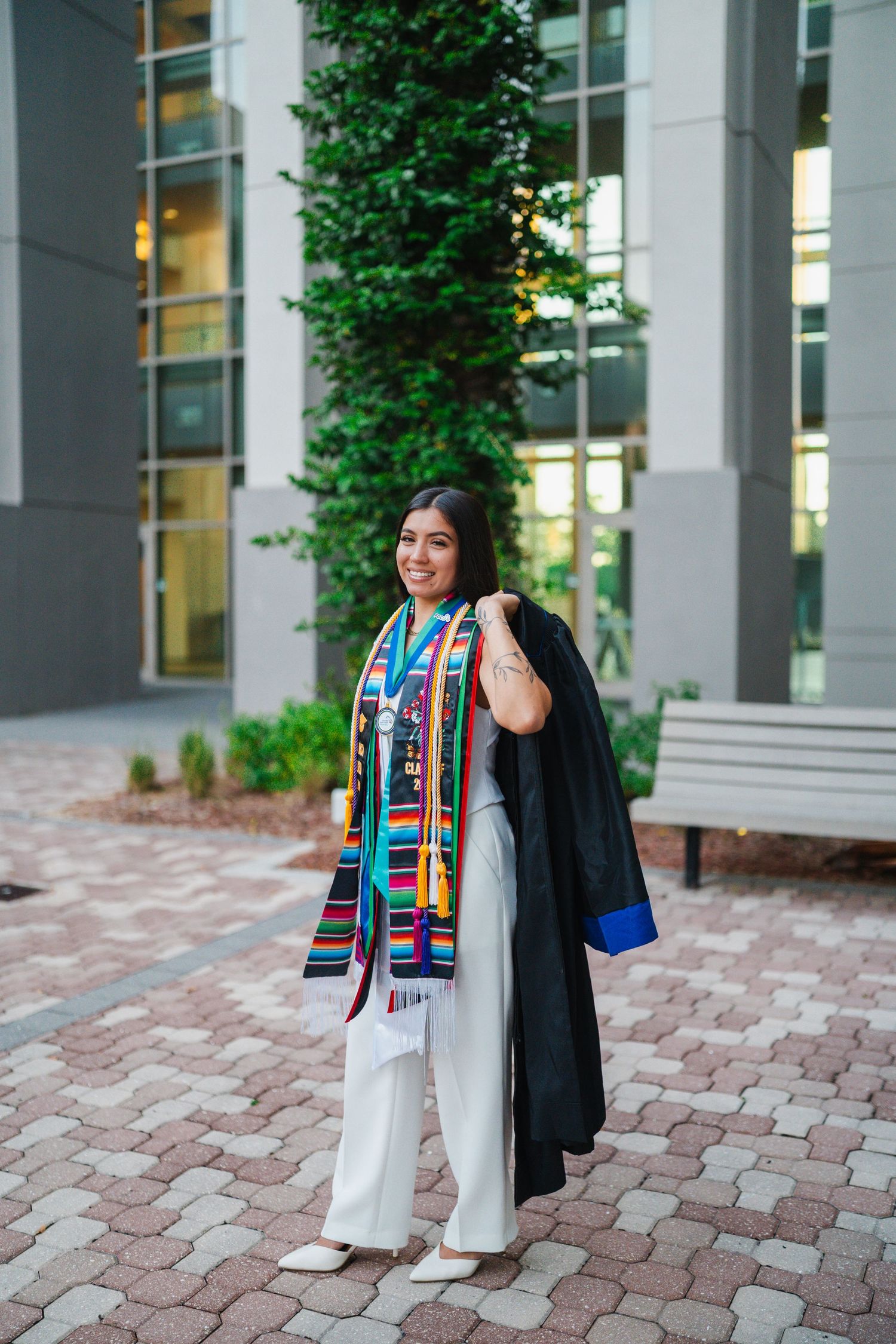 Smiling female graduate in white outfit holding black gown, wearing colorful stole, posing outdoors near modern building.