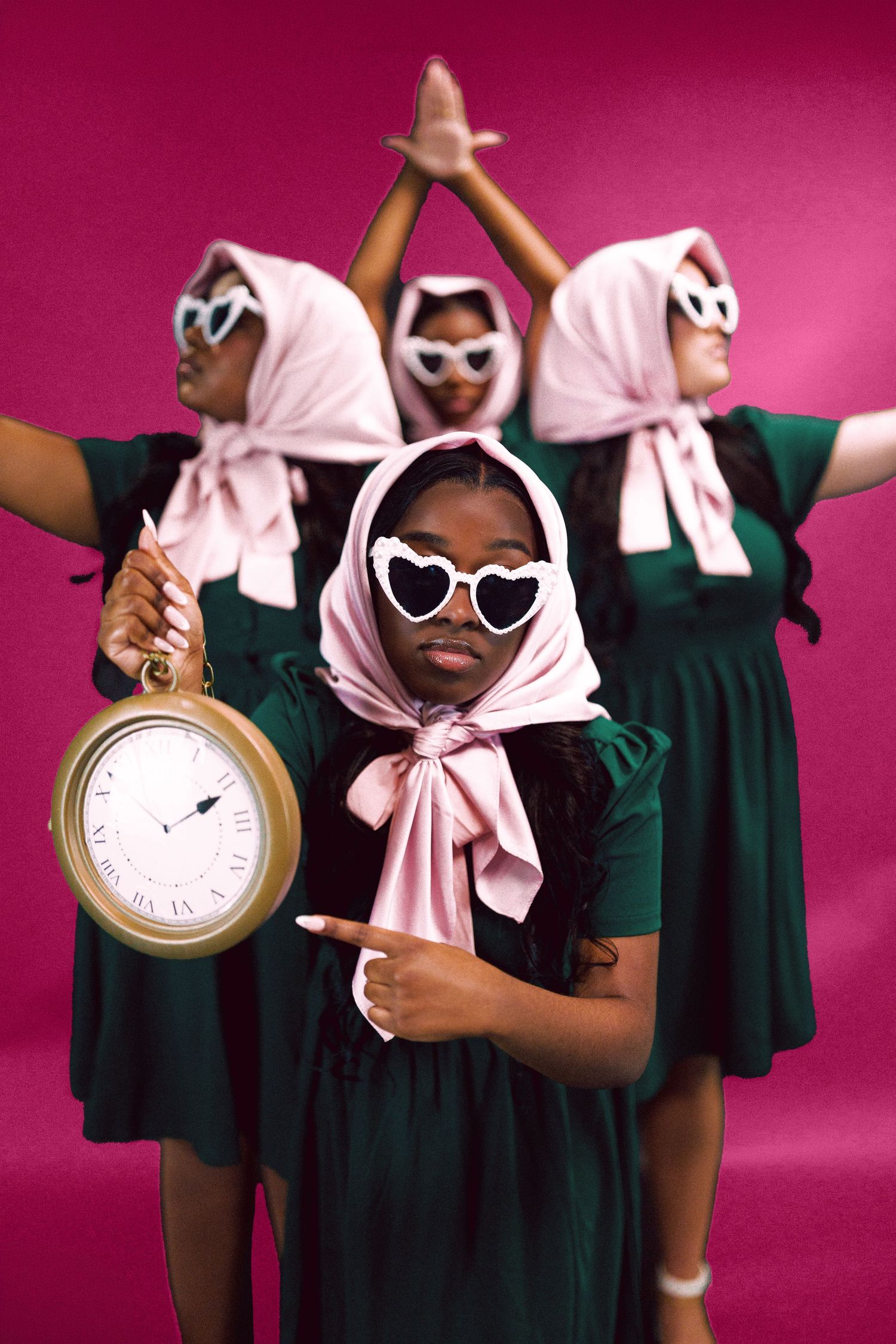 Three women in green dresses, pink headscarves, and heart sunglasses pose with a large clock on a pink background.