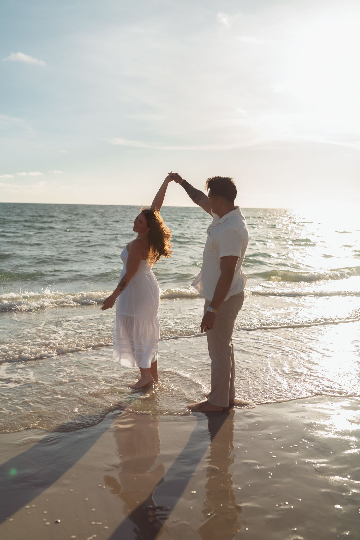 Romantic couple dancing on a sunlit beach, woman in white dress, waves gently lapping at their feet.