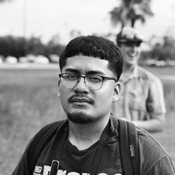 Black and white portrait of person wearing glasses and t-shirt standing outdoors with blurred figure in background.