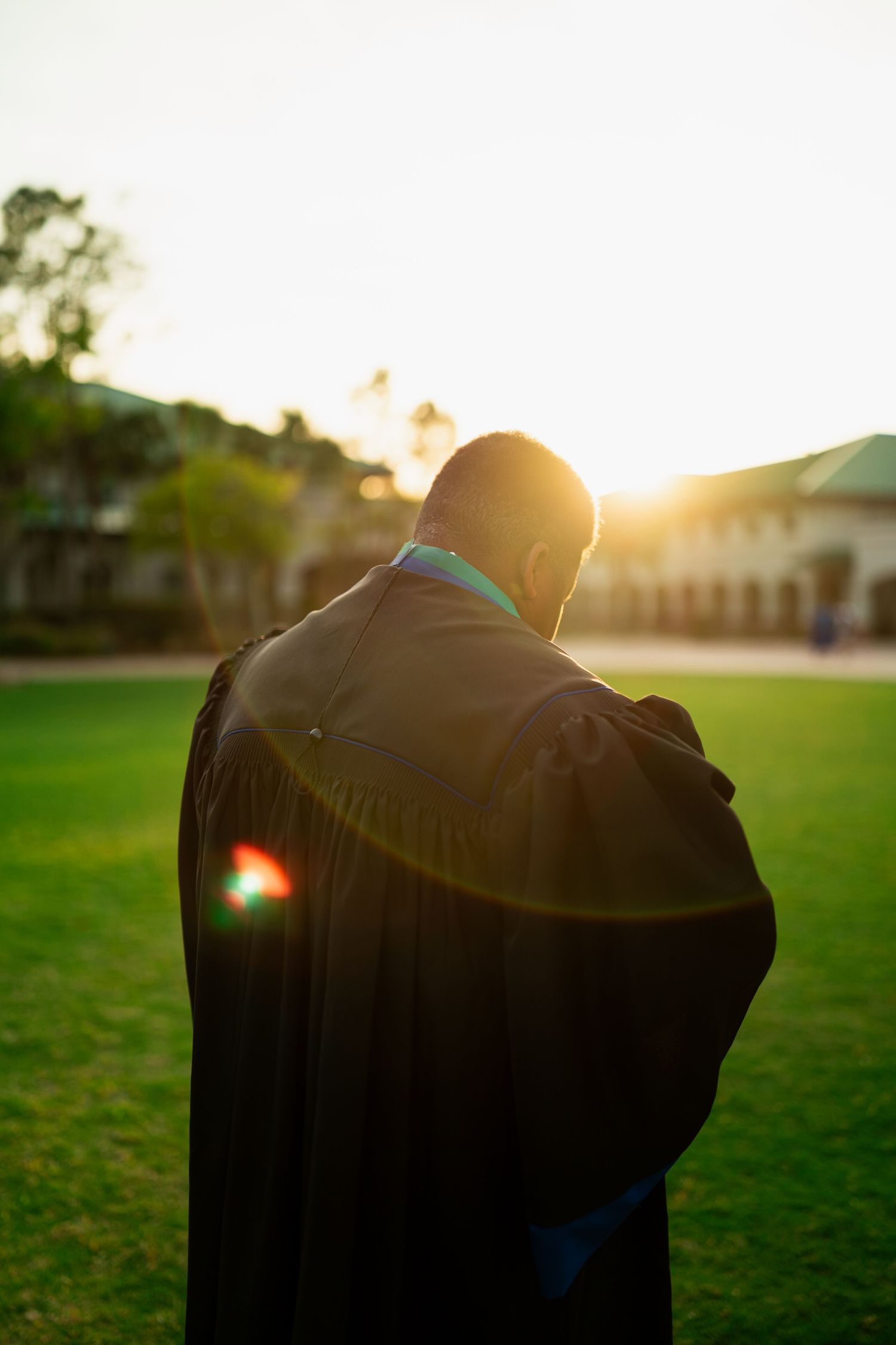 Graduate in black gown stands on green lawn at sunset with warm golden light glowing behind them.