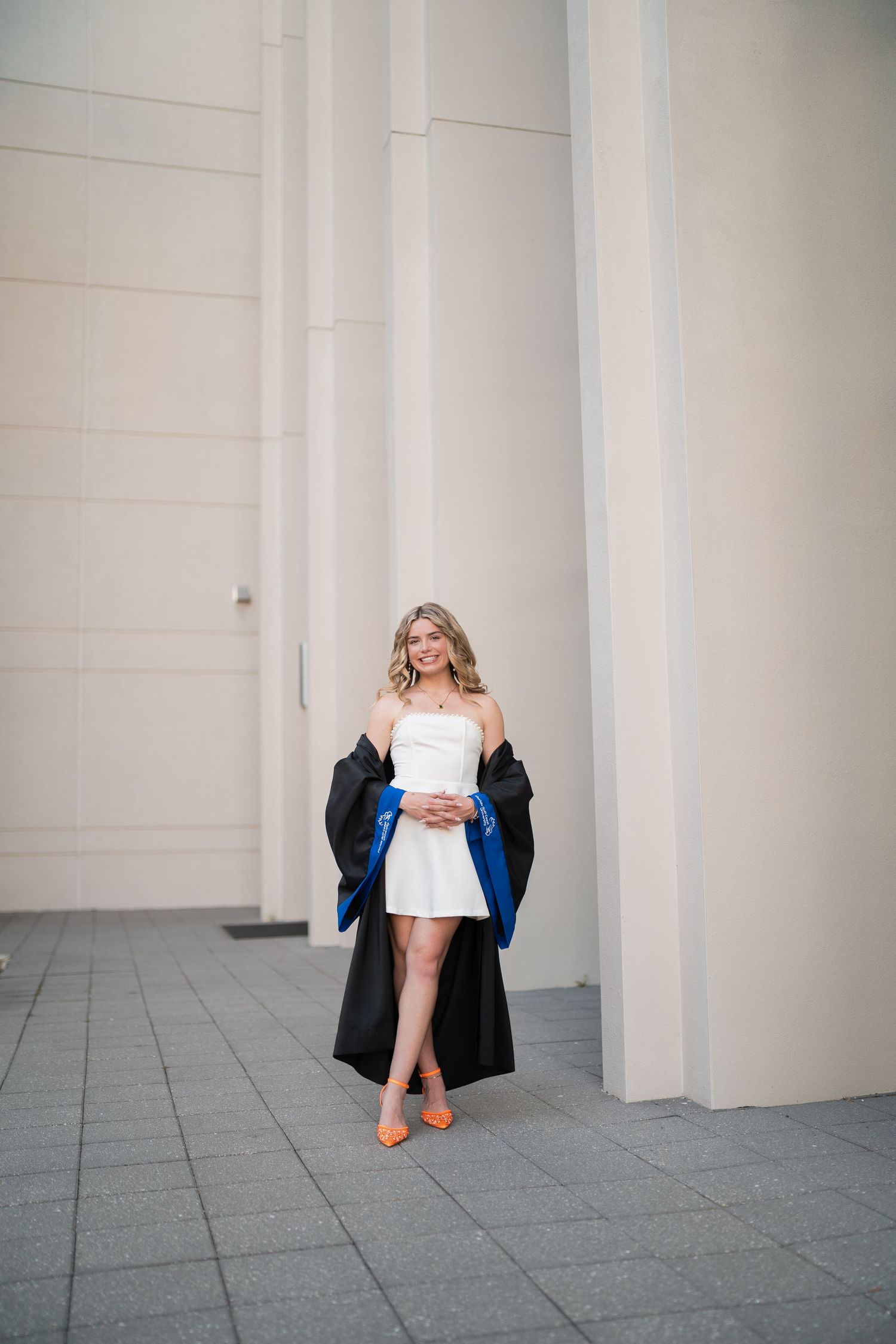 Young woman in white dress and graduation gown with blue hood smiling outdoors near modern building columns.