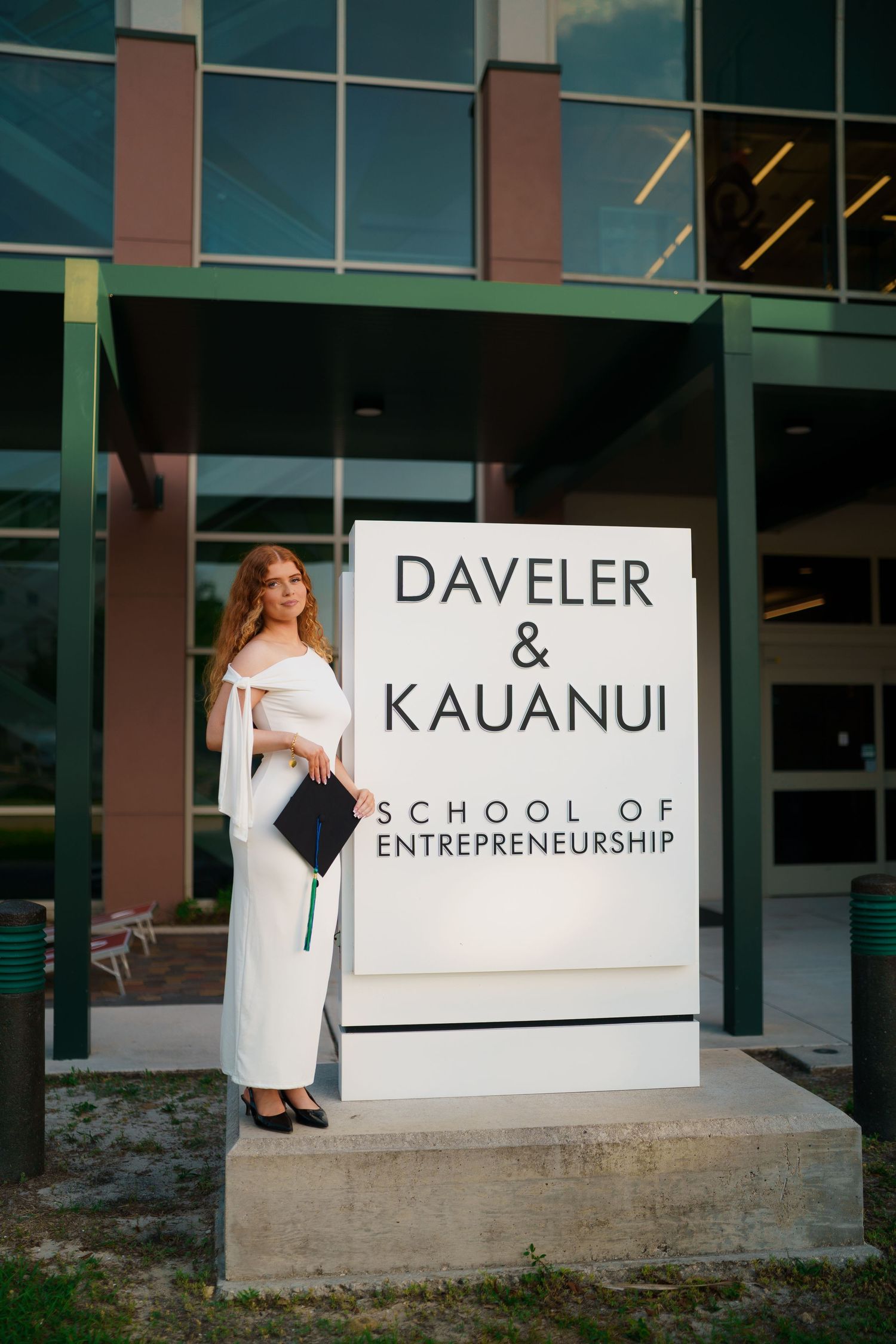 Graduate in white dress holds cap beside Daveler & Kauanui School of Entrepreneurship sign at university building.