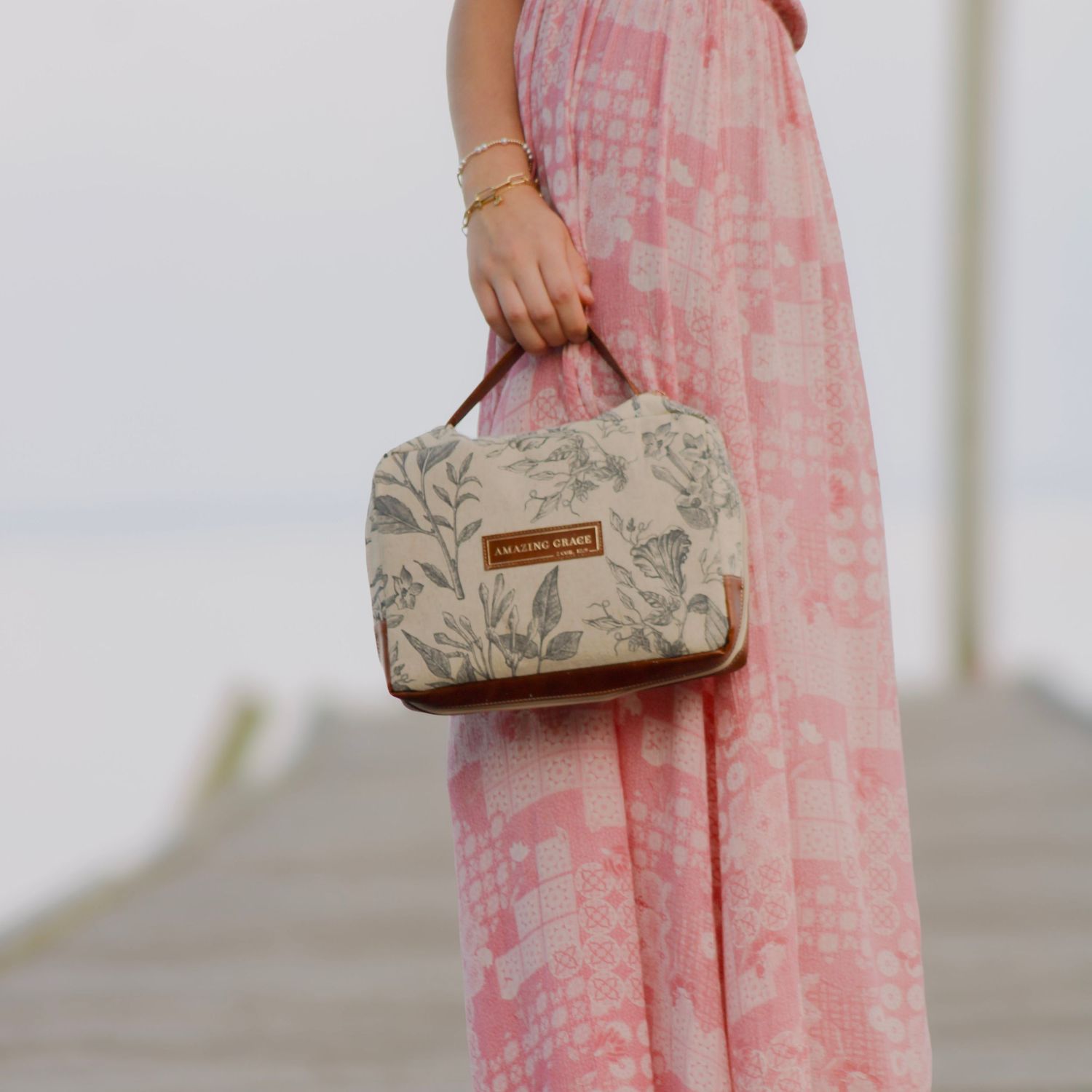 Woman holding a floral patterned cosmetic bag while wearing a flowing pink floral maxi dress on a wooden dock.