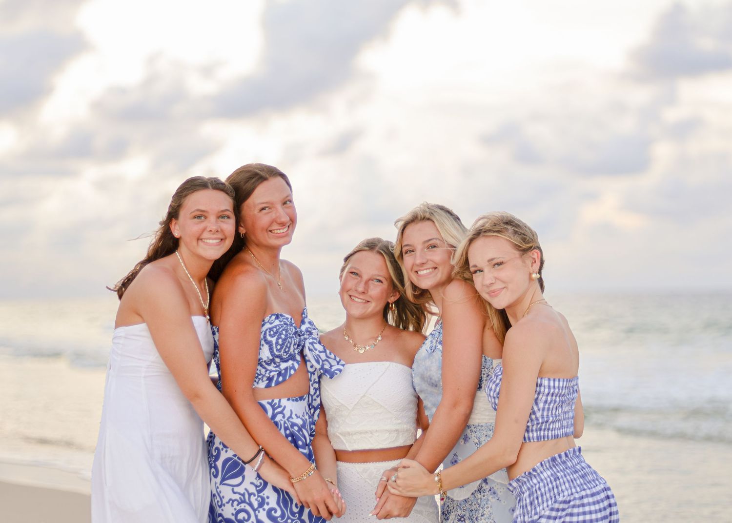 Group of women in blue and white dresses pose together on a beach at sunset.