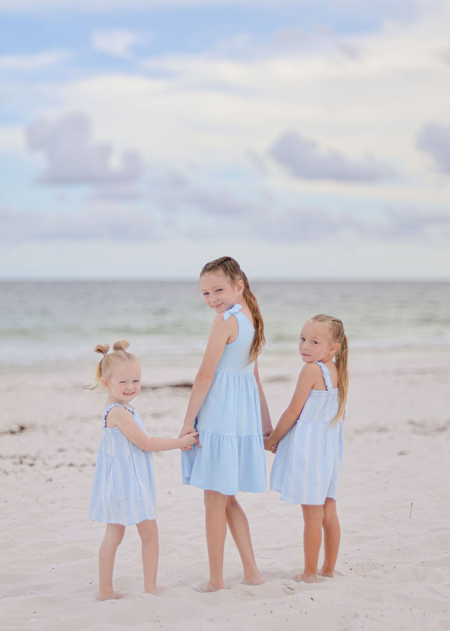 Three young girls in matching light blue dresses holding hands while standing on a beach at sunset.