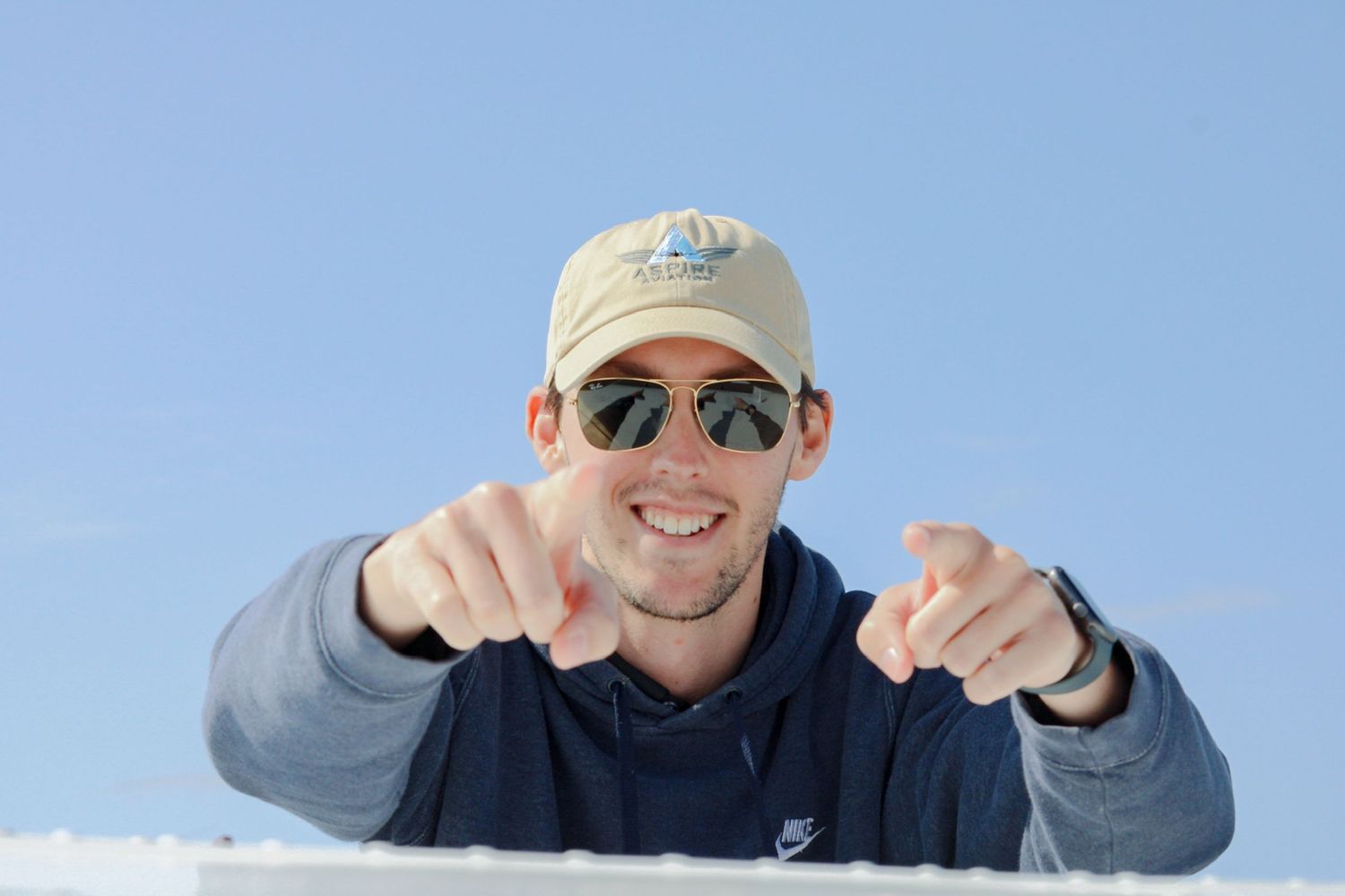 Person in a baseball cap and sunglasses pointing forward against blue sky.