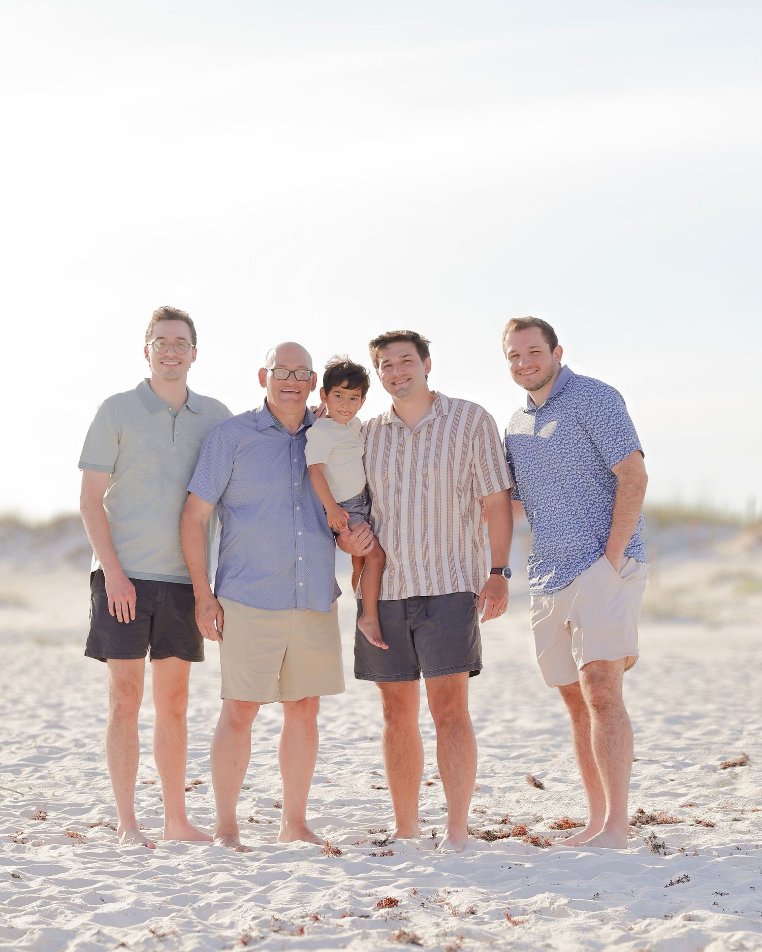 A family group stands together on a sandy white beach during golden hour wearing casual summer clothing.