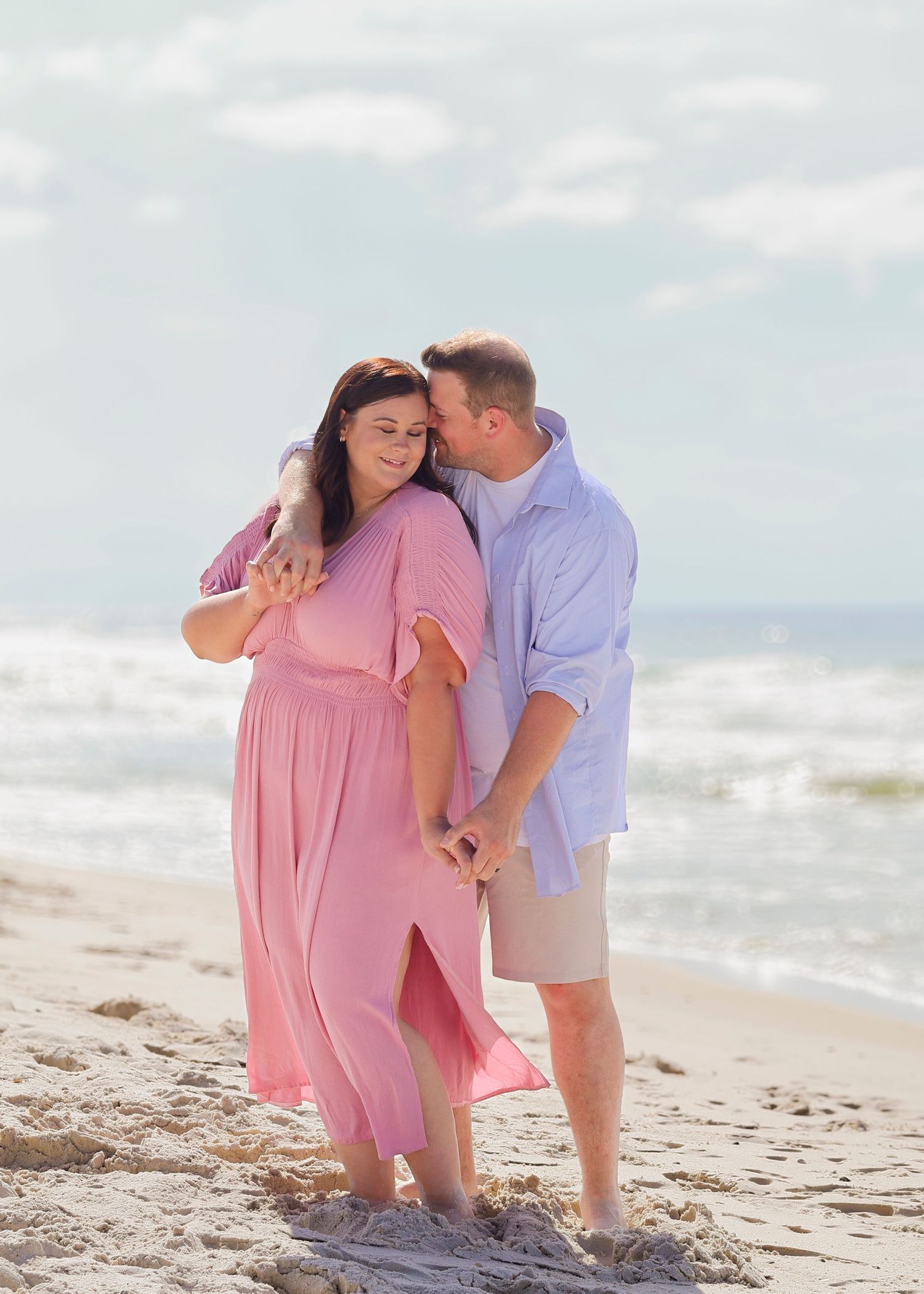 Romantic couple embraces on a sunny beach, with person in pink maxi dress and partner in light blue shirt and khaki shorts.