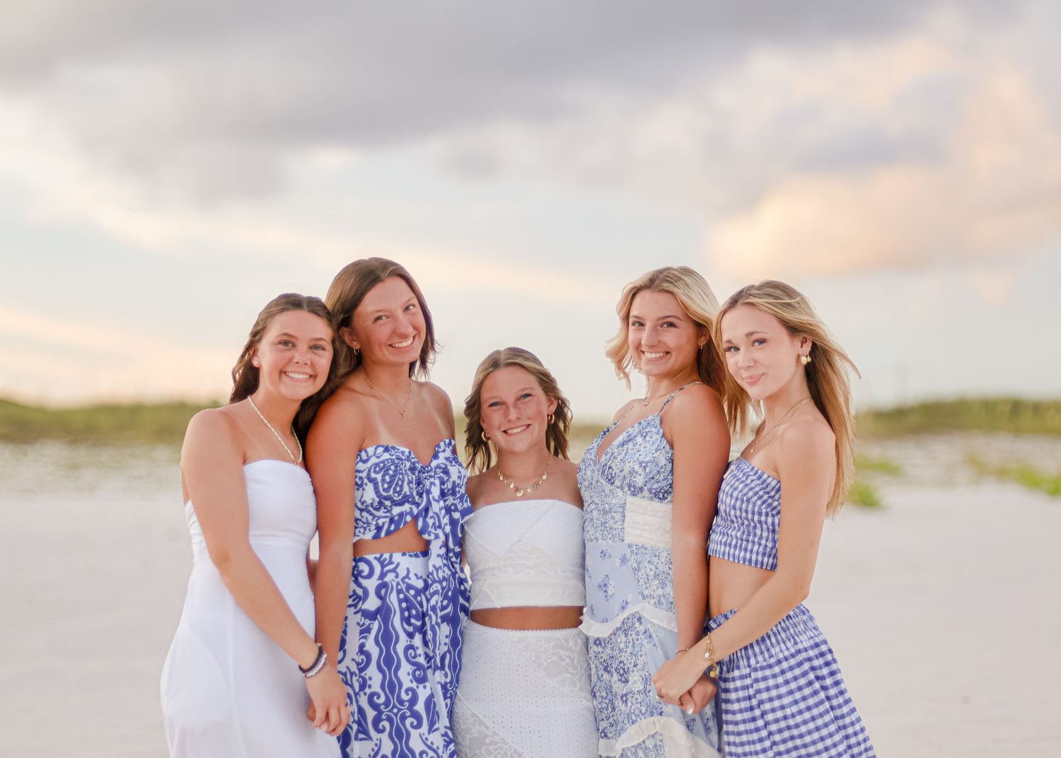 A group of people wearing blue and white summer dresses pose together on a sandy beach at sunset.