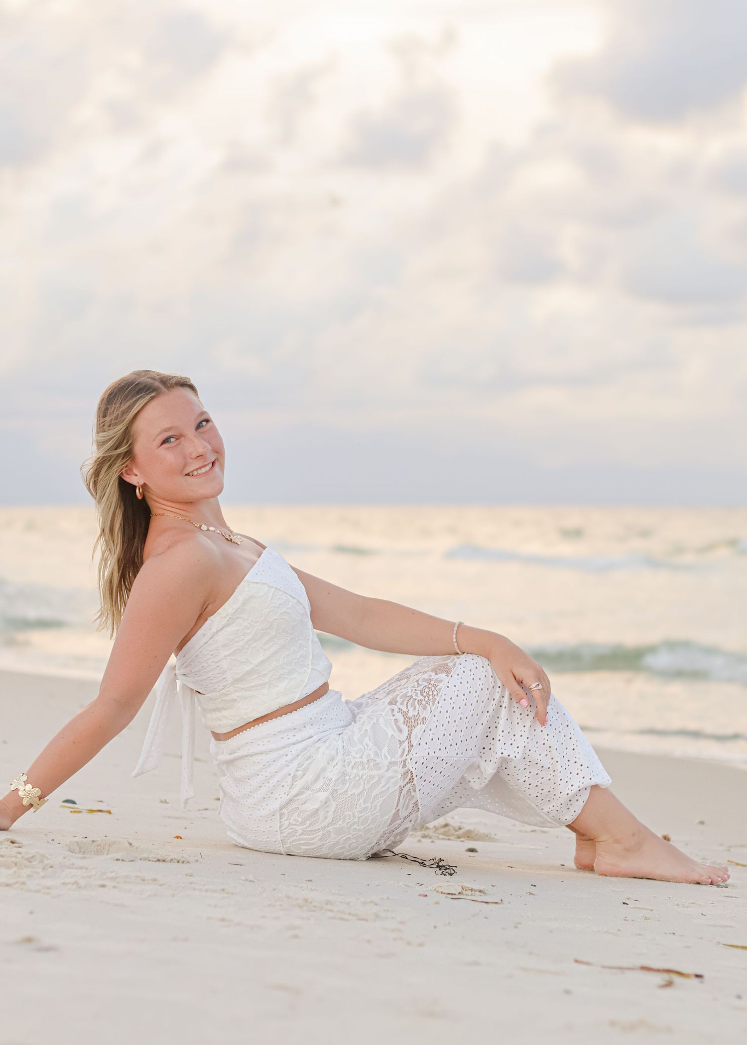 Woman in white lace outfit relaxing on sandy beach at sunset with gentle waves and cloudy sky in background.