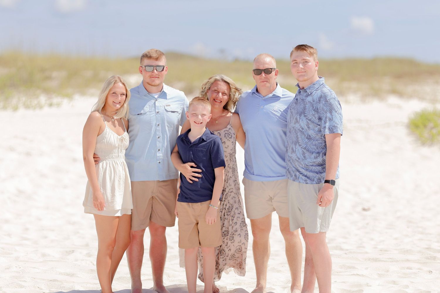 Group stands together smiling on a sunny white sand beach with dunes in the background on a clear day.
