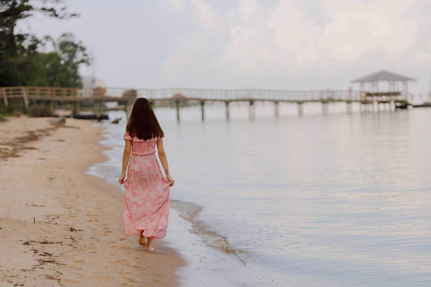 Someone in a flowing pink dress walks along a peaceful beach with a wooden pier stretching into calm waters at sunset.