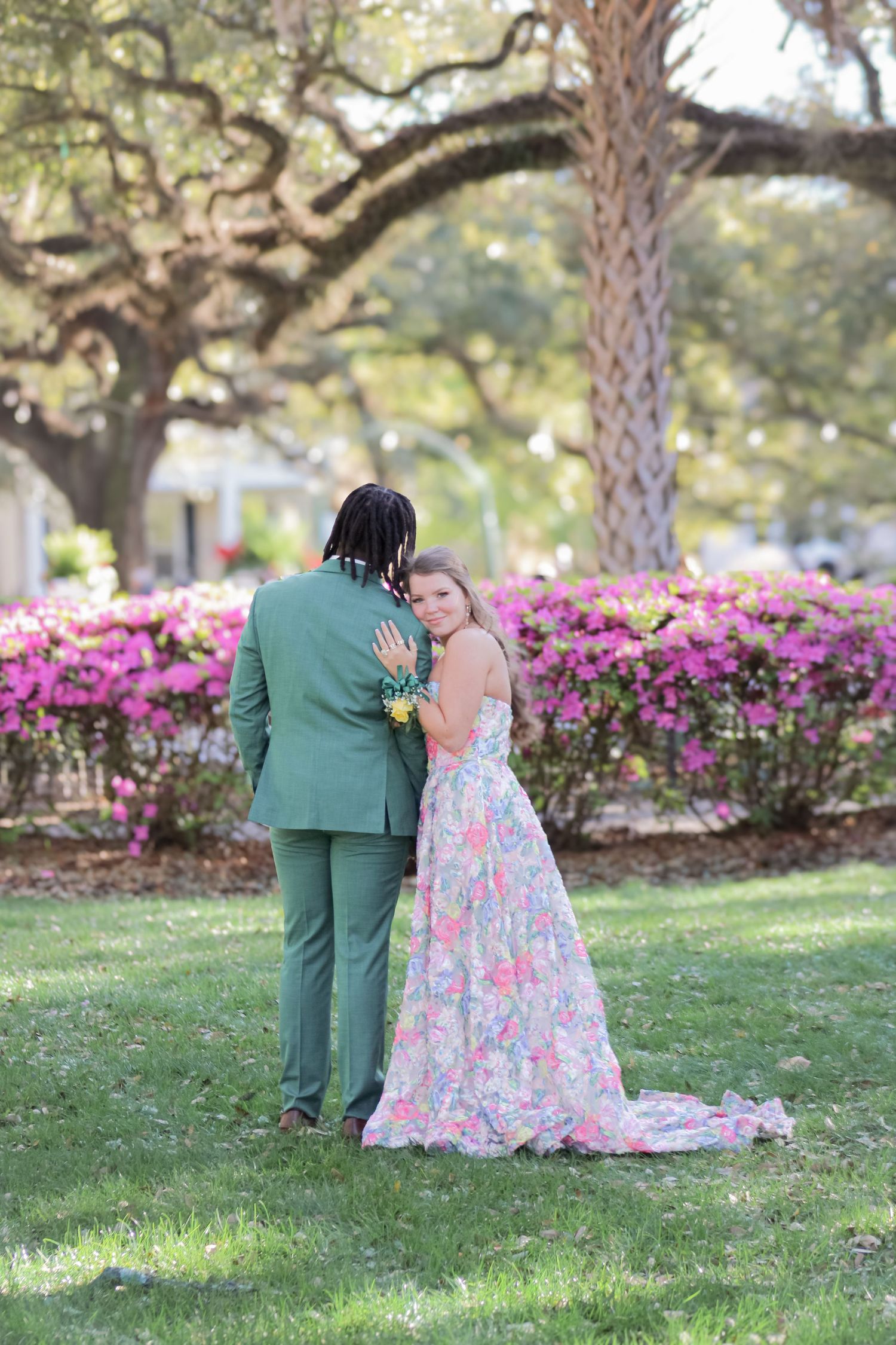 A couple shares an intimate moment in a garden with blooming azaleas and oak trees in the background.