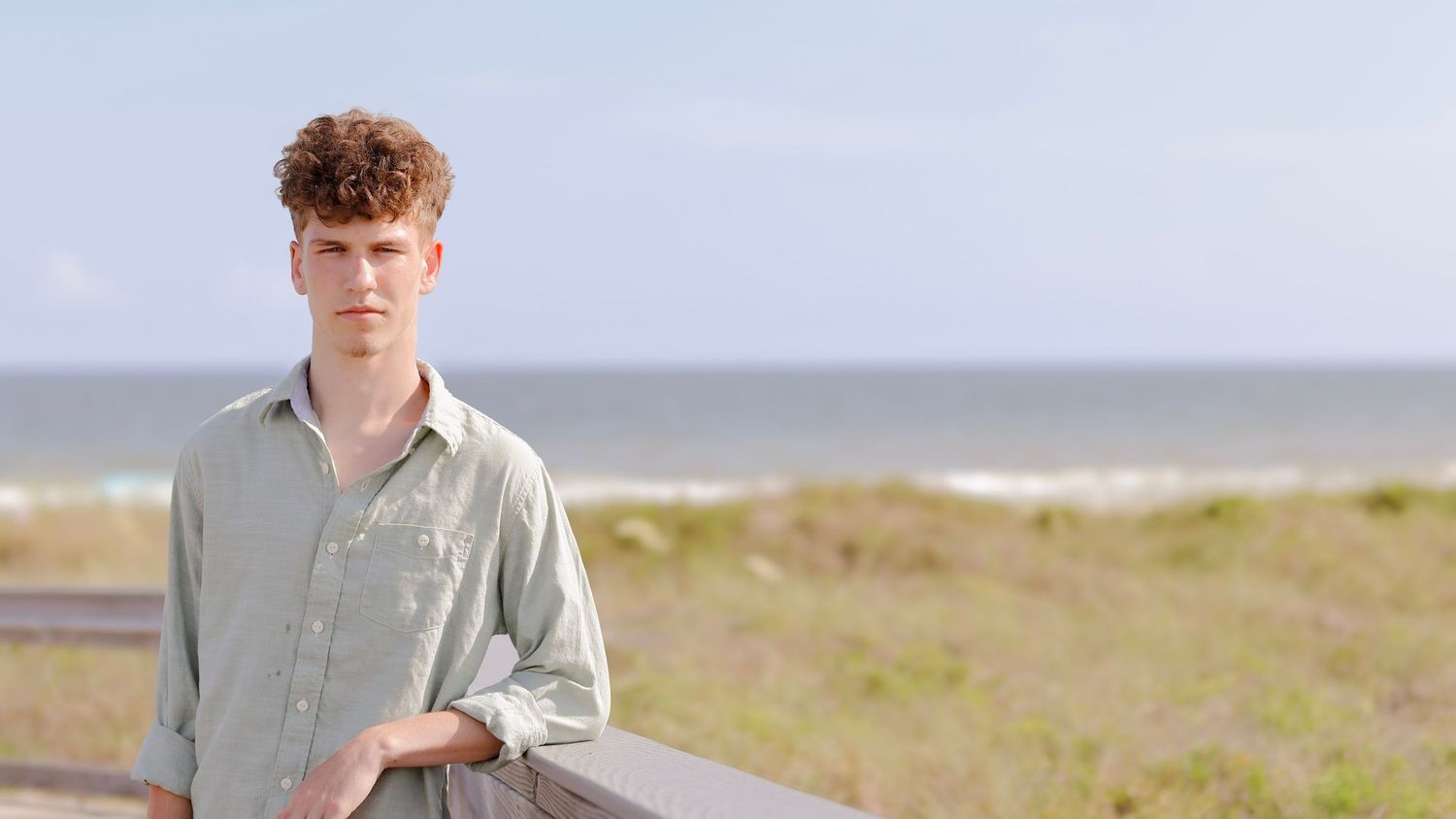 A man walks along a scenic coastal path with beach grass and ocean views on a cloudy day.