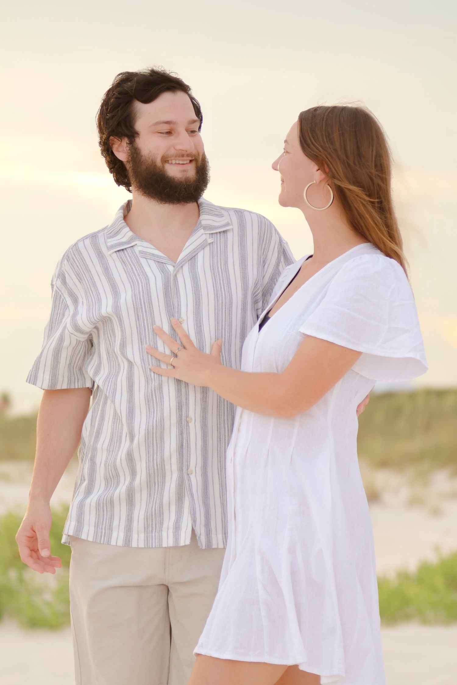 A couple laughs and enjoys a romantic moment together on a sun-lit beach wearing white and beige clothing.