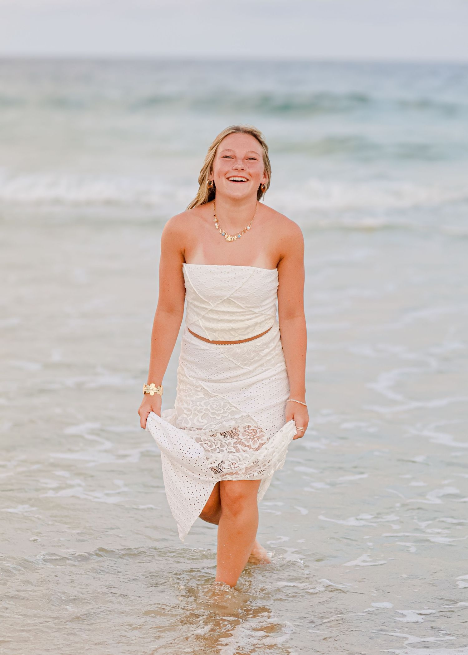 Someone in a white lace dress laughs joyfully while standing in shallow ocean waves at the beach.