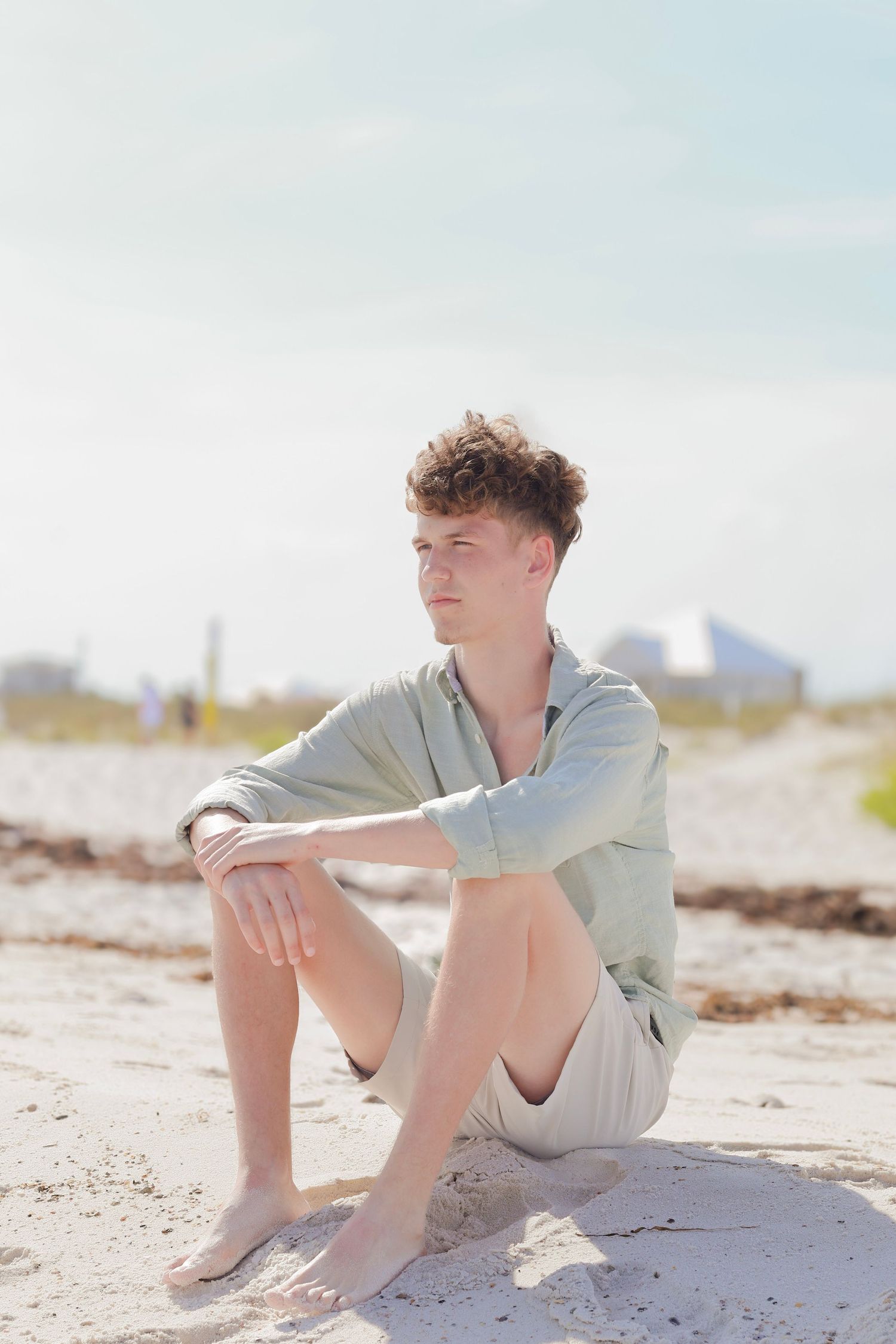 A person in a light gray hoodie sits contemplatively on a sandy beach on a bright sunny day.