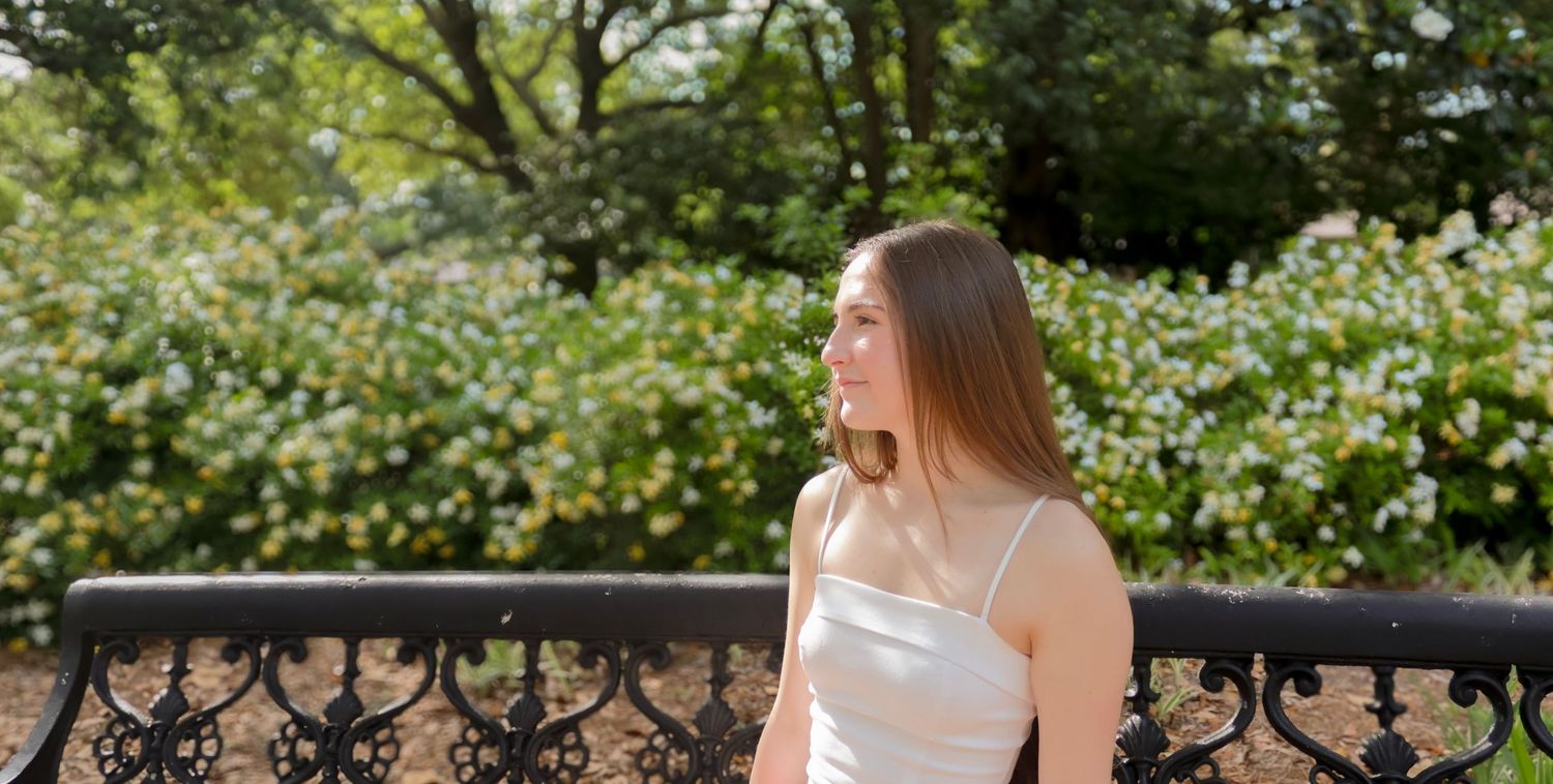 Young woman in white dress standing on balcony with flowering bushes and trees in background on sunny day.