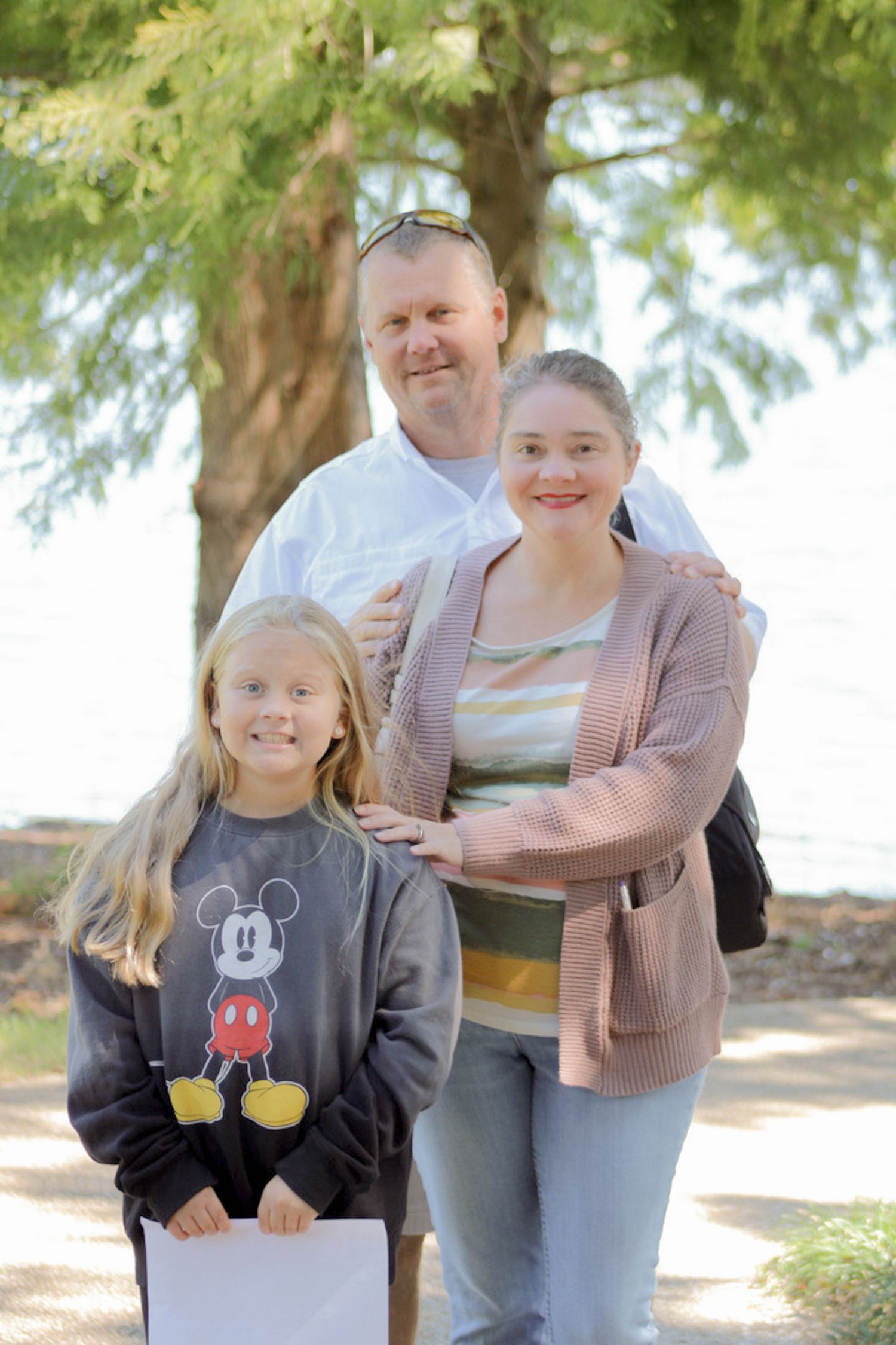 A small family poses together outdoors under trees on a sunny day, with one child wearing a Mickey Mouse sweater.