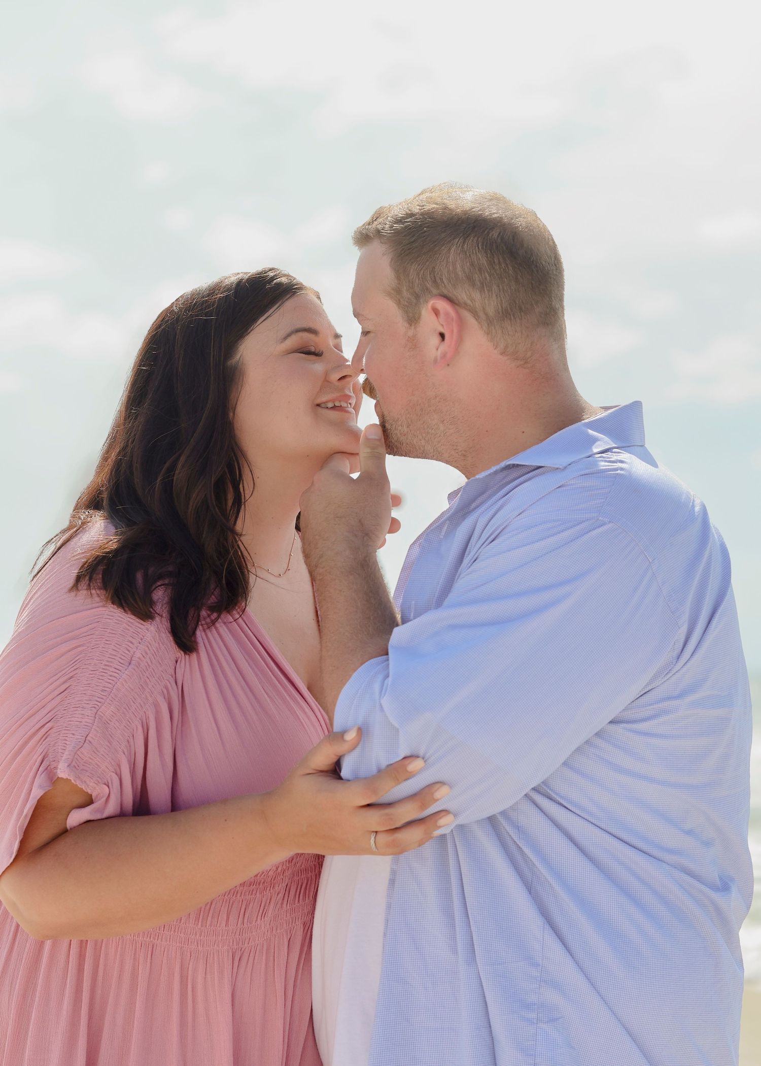 A loving couple shares a joyful moment together, with her wearing pink and him in light blue against a soft sky background.