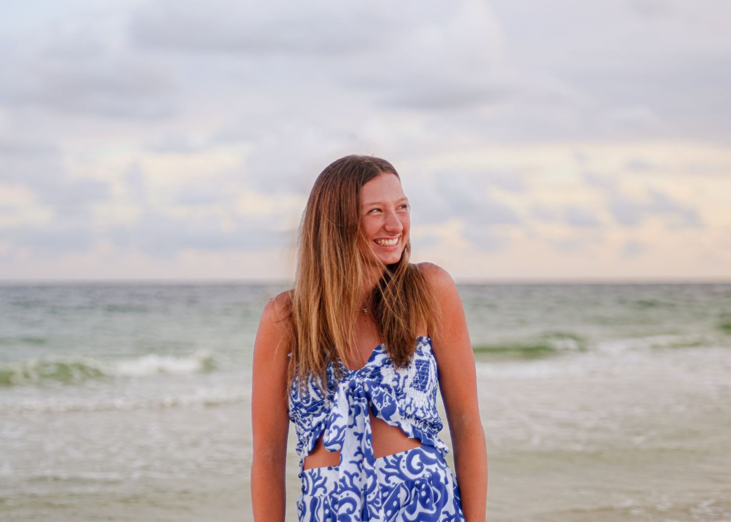 A person in a blue and white floral dress standing on a beach at sunset with waves in the background.