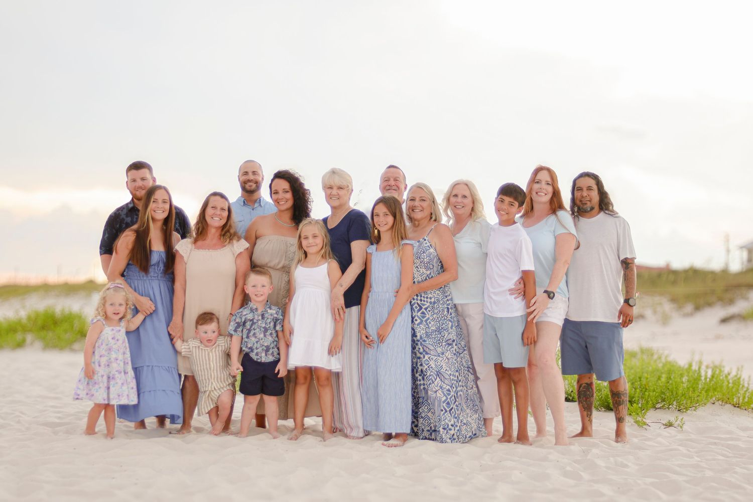 A group in coordinated white and blue outfits poses together for a family portrait on the beach at sunset.
