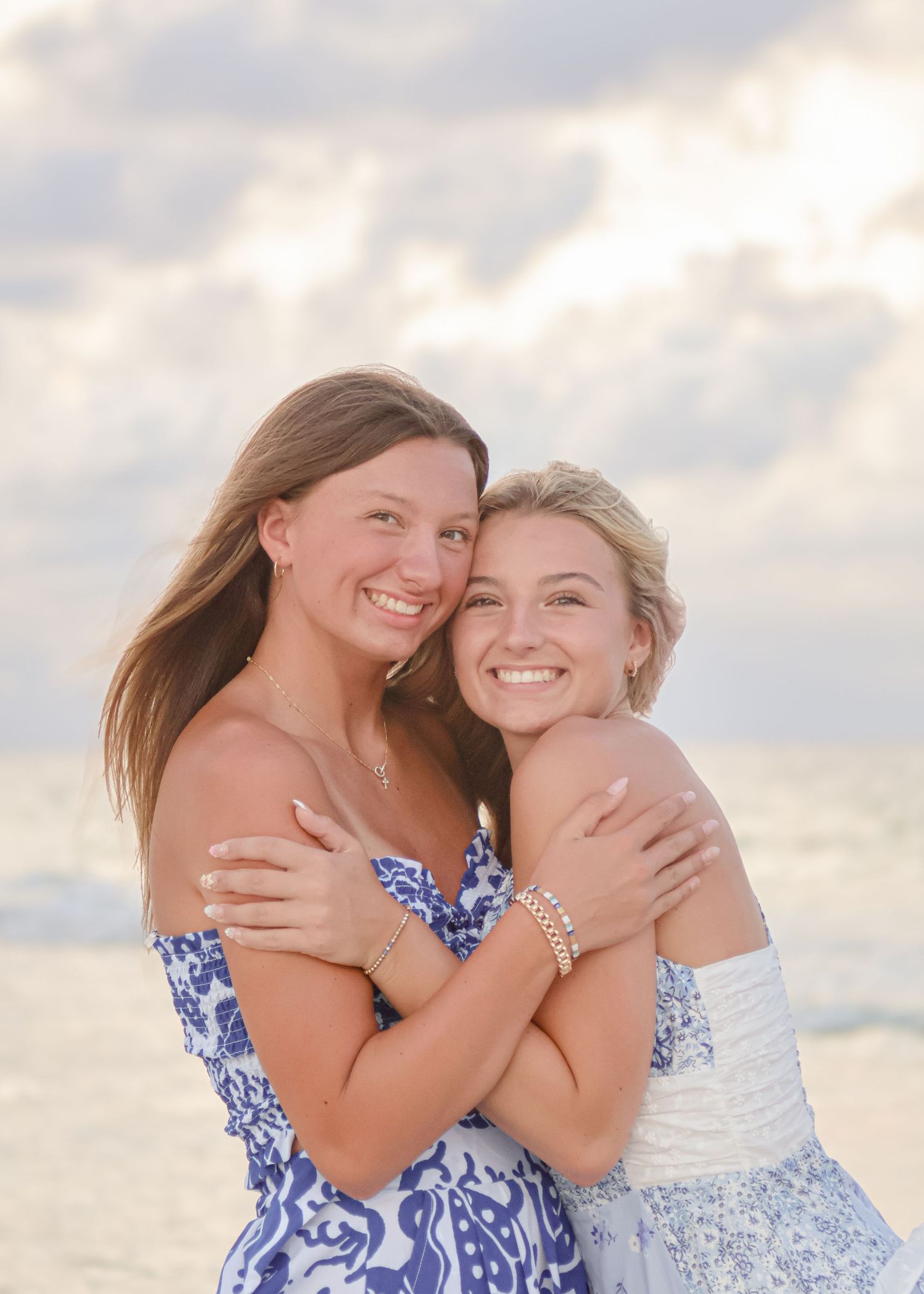 Two friends in blue and white dresses embrace and smile while posing together on a beach at sunset.