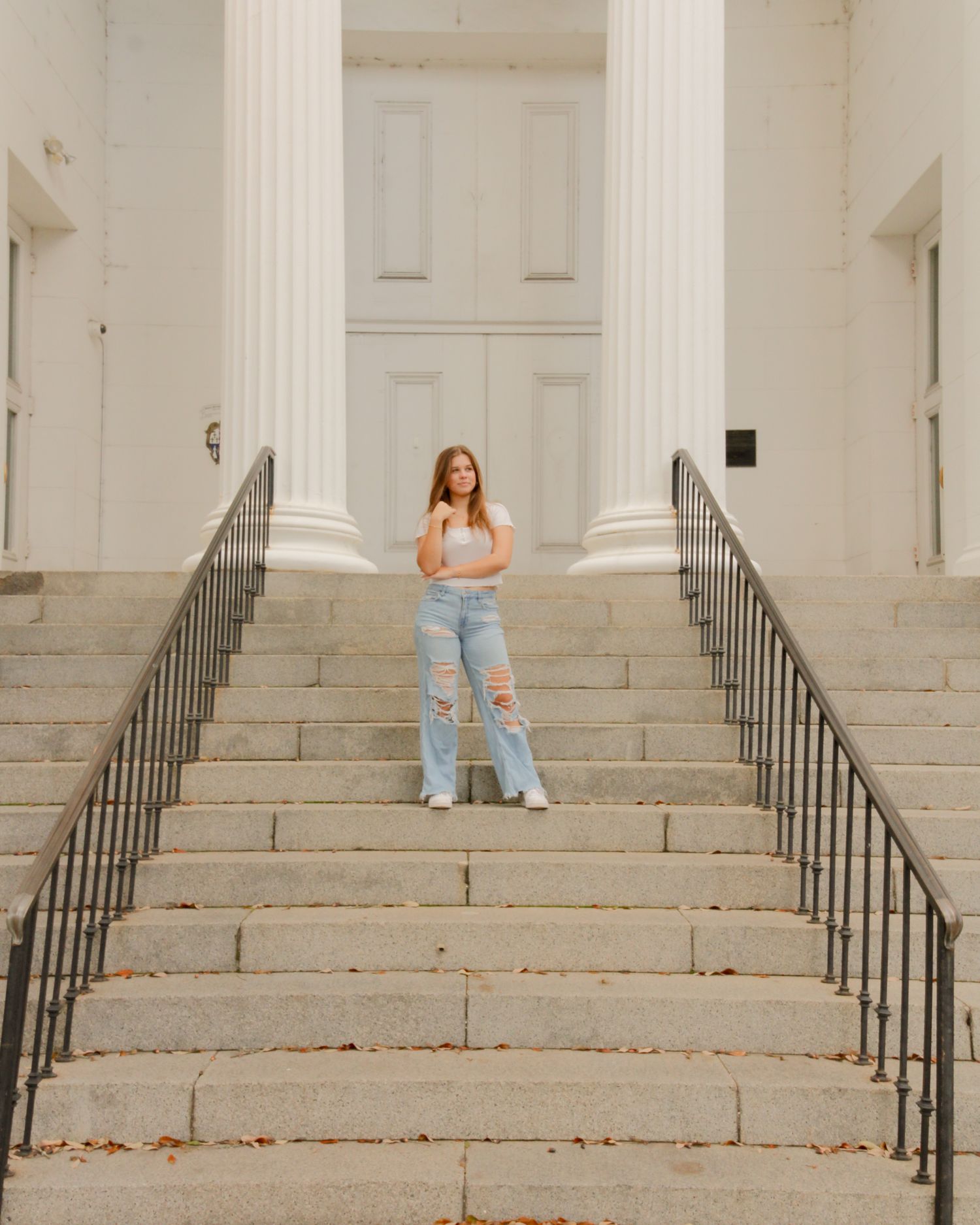 Person in light blue jeans and white top poses on grand stone staircase with white columns in background.