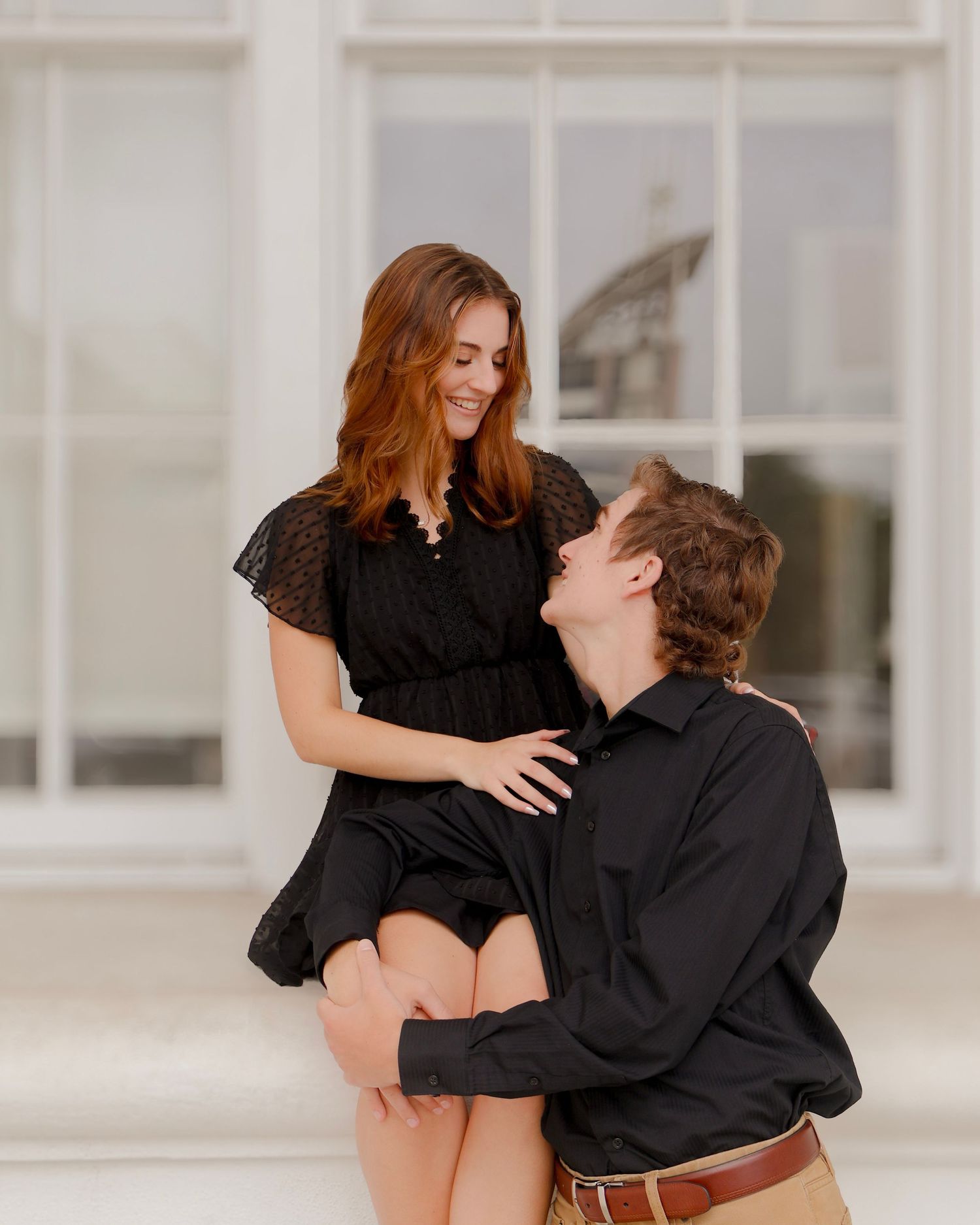 A tender romantic moment shared between a couple in matching black outfits sitting by a bright window.