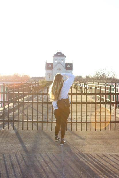 Figure in white shirt and brown pants stands at iron fence overlooking church building during golden sunset.