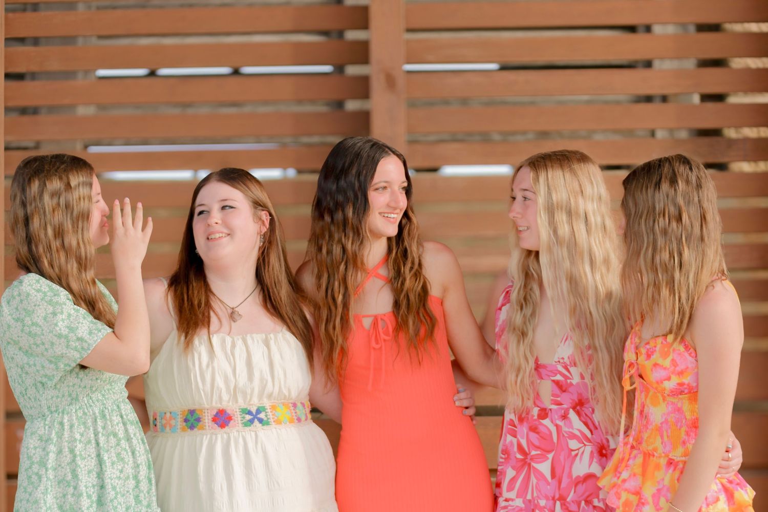A group of five women in colorful spring dresses laugh and chat together in front of a wooden wall.