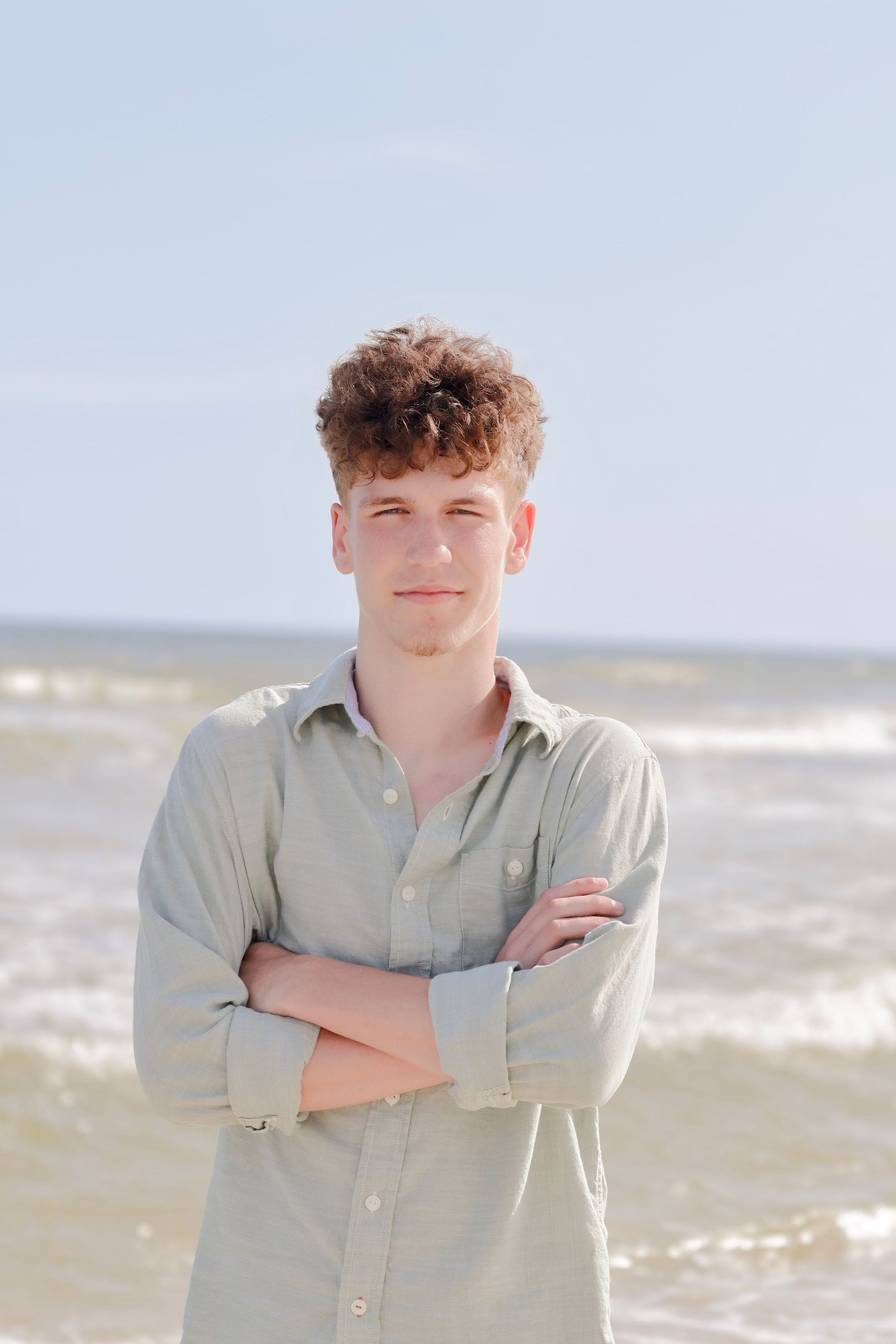 A person in a light button-up shirt stands with crossed arms on a sunny beach with waves rolling in behind them.