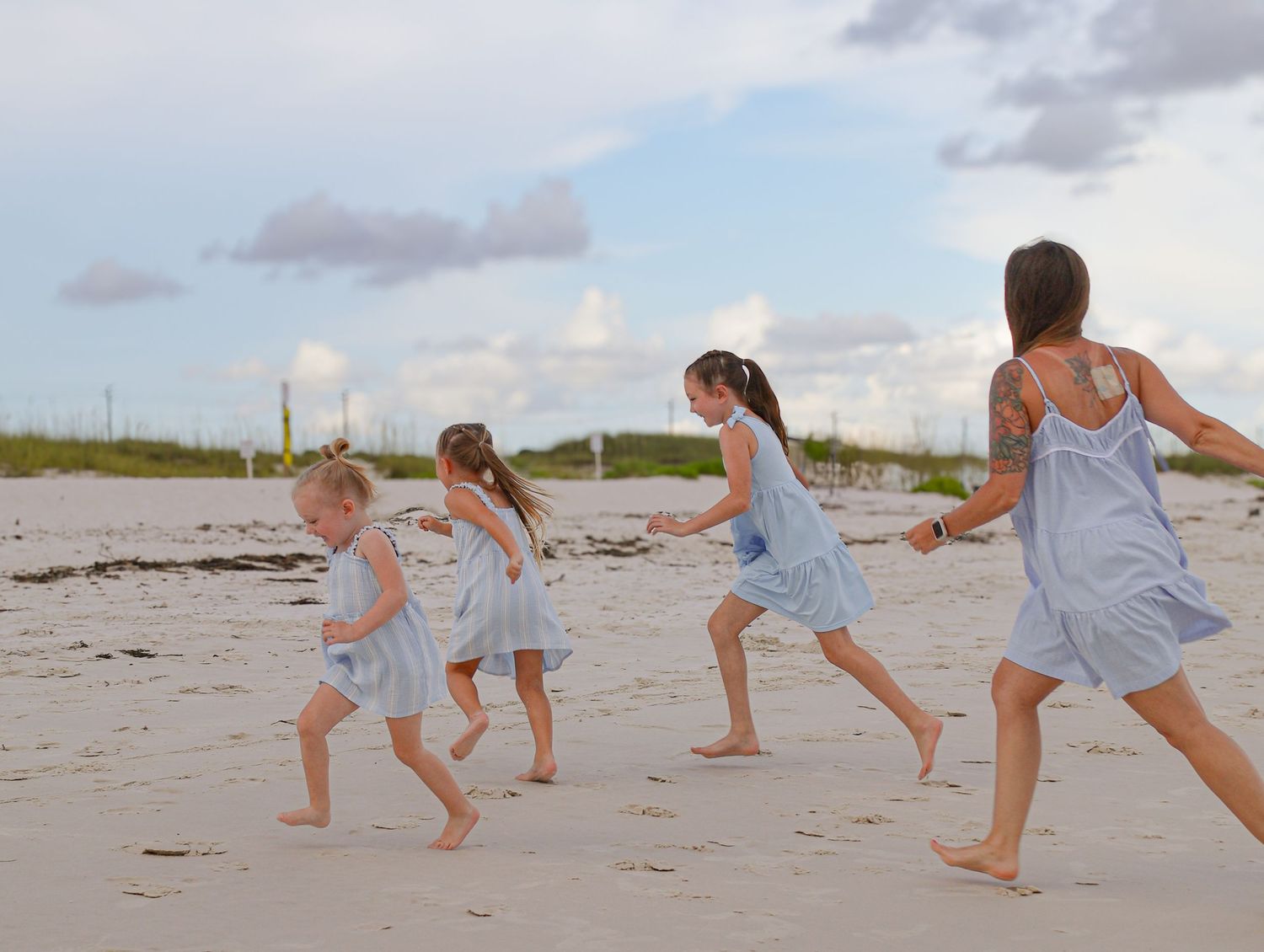 A group wearing matching light blue dresses runs playfully along a sandy beach at sunset.