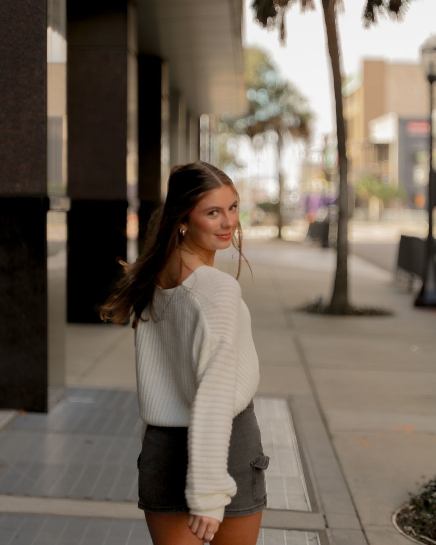 A person in a white sweater and dark shorts looking back over their shoulder while walking down a palm tree-lined street.