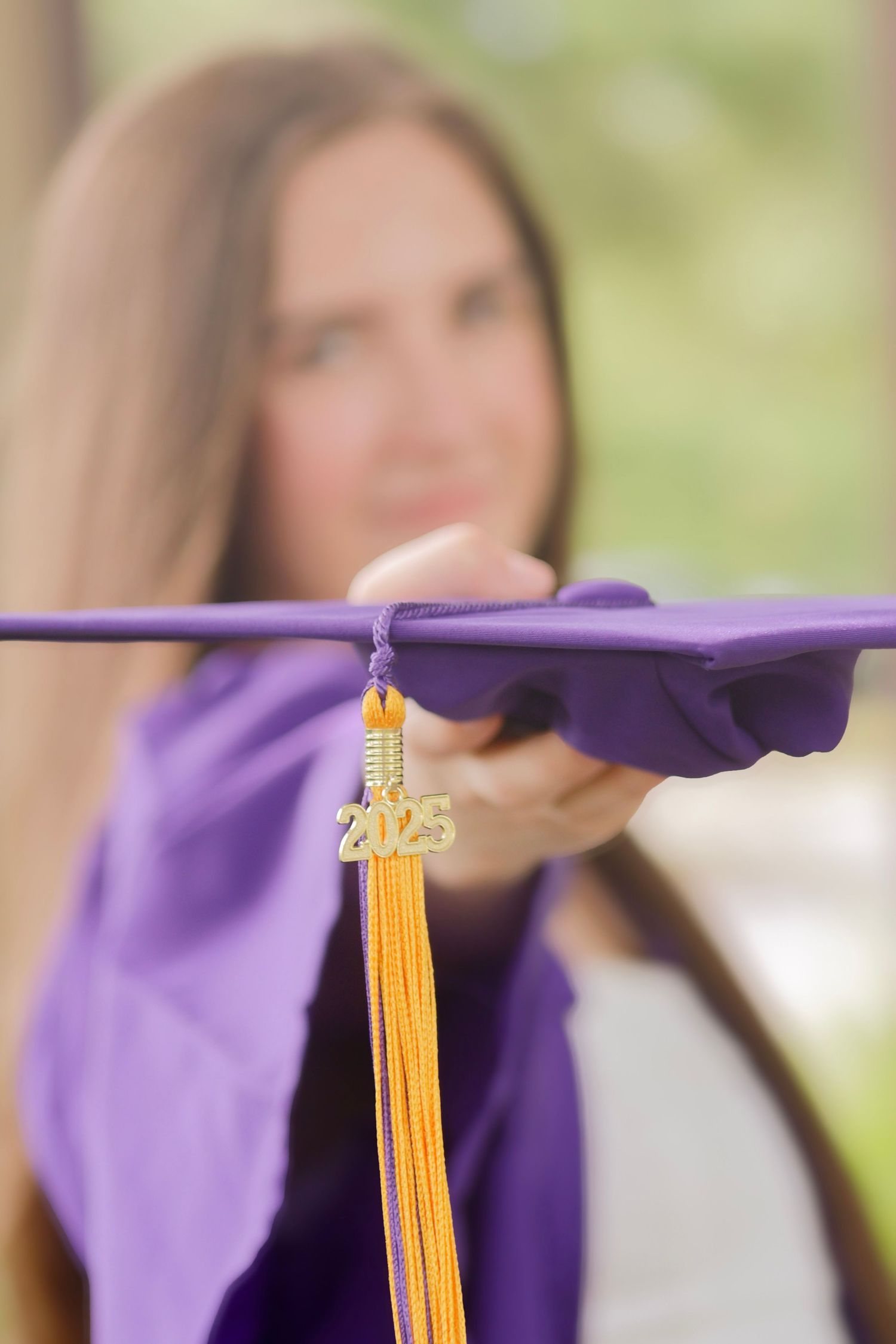 A graduation cap with gold 2023 tassel in focus against a blurred purple gown and natural background.